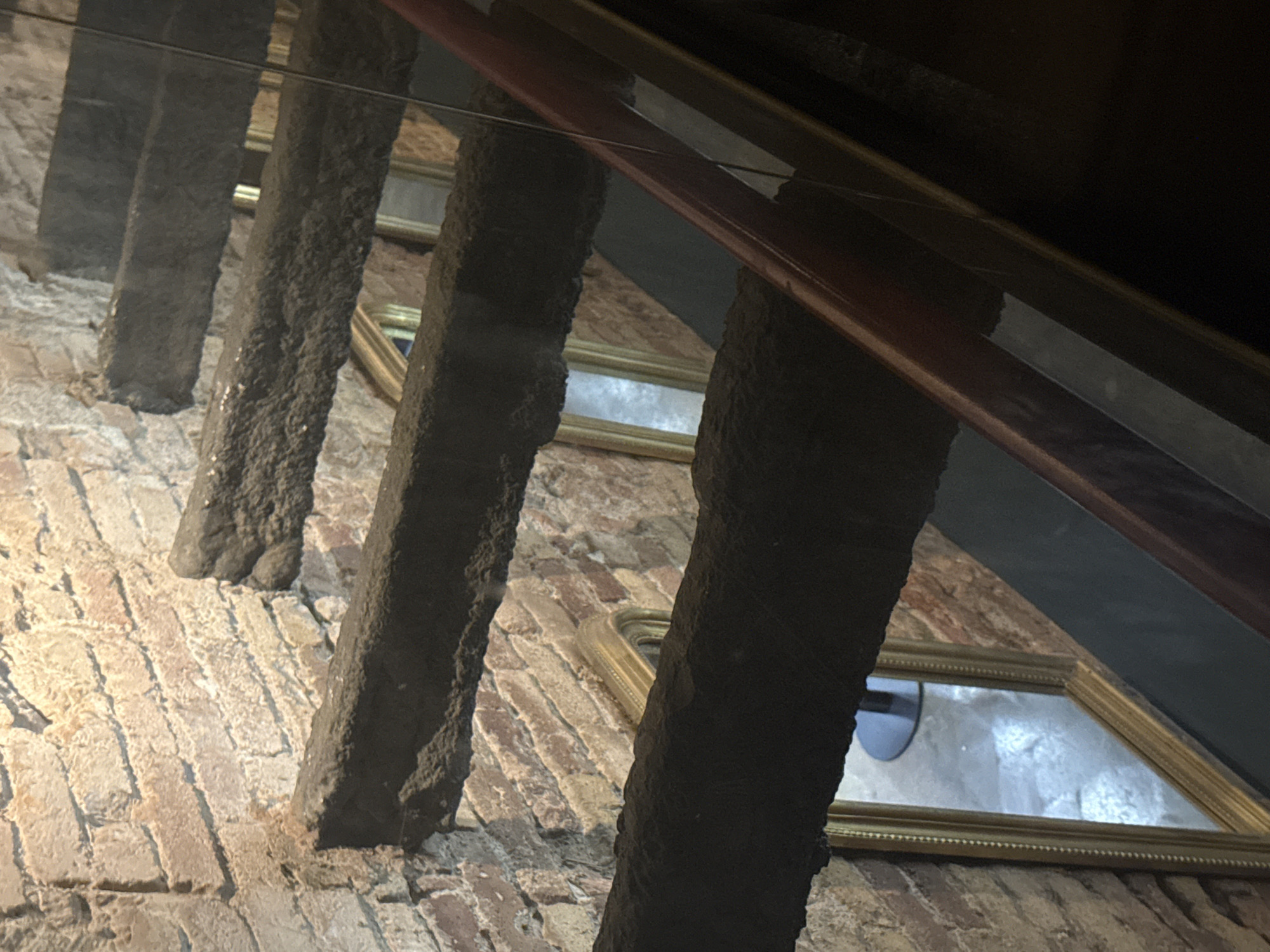 This image shows a view looking down a staircase made of rough-hewn stone steps supported by brick walls. Several ornate, gold-framed mirrors are visible between the steps, reflecting the surrounding brickwork and the steps themselves. A dark red handrail runs along the right side of the image, separated from the staircase by a glass barrier. The overall impression is of an old, possibly historic, interior. The scene is likely located within the Gothic Quarter of Barcelona, Spain, known for its narrow streets and ancient architecture, often featuring similar exposed brick and reflective surfaces in its historical buildings.