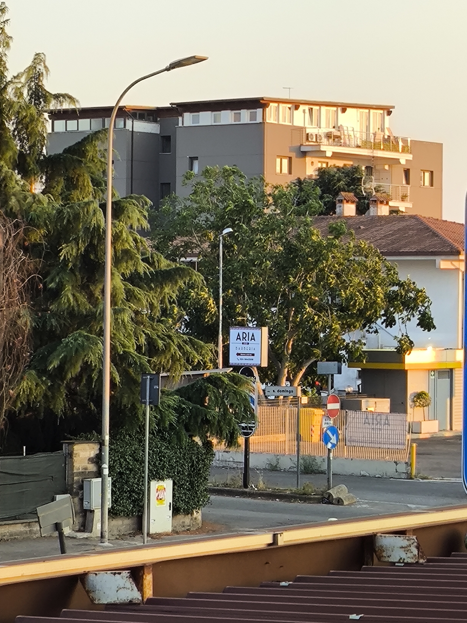 The image depicts an outdoor scene in a city, likely during the daytime, given the lighting. In the foreground, a corrugated metal surface with visible rivets and rust suggests a rooftop or barrier. Behind this, a street and sidewalk are visible.
Several trees with dense green foliage are prominent, with one particularly large tree in the mid-ground. A tall lamppost stands on the left side of the image, and another on the right, though the right one is partially obscured.
Various buildings are present. A multi-story building with a gray facade, white balconies, and numerous windows is in the background. To the right of this, a smaller, lighter-colored building with a pitched roof is visible, featuring several chimneys. Further to the right, a white building with a yellow awning is partially visible.
Street furniture includes traffic signs, a utility box on a pole, and a small, light-colored structure with a door that appears to be a kiosk or small building. Several smaller electrical boxes are also mounted on poles. A sign with the word "ARIA" and "BARBERIA" is mounted on a pole near the large tree, indicating a business. Other street signs, including a circular red and white sign and a blue directional arrow sign, are also present.
The ground is paved, with some areas of grass or vegetation visible. The overall impression is of an urban streetscape with residential and commercial elements.