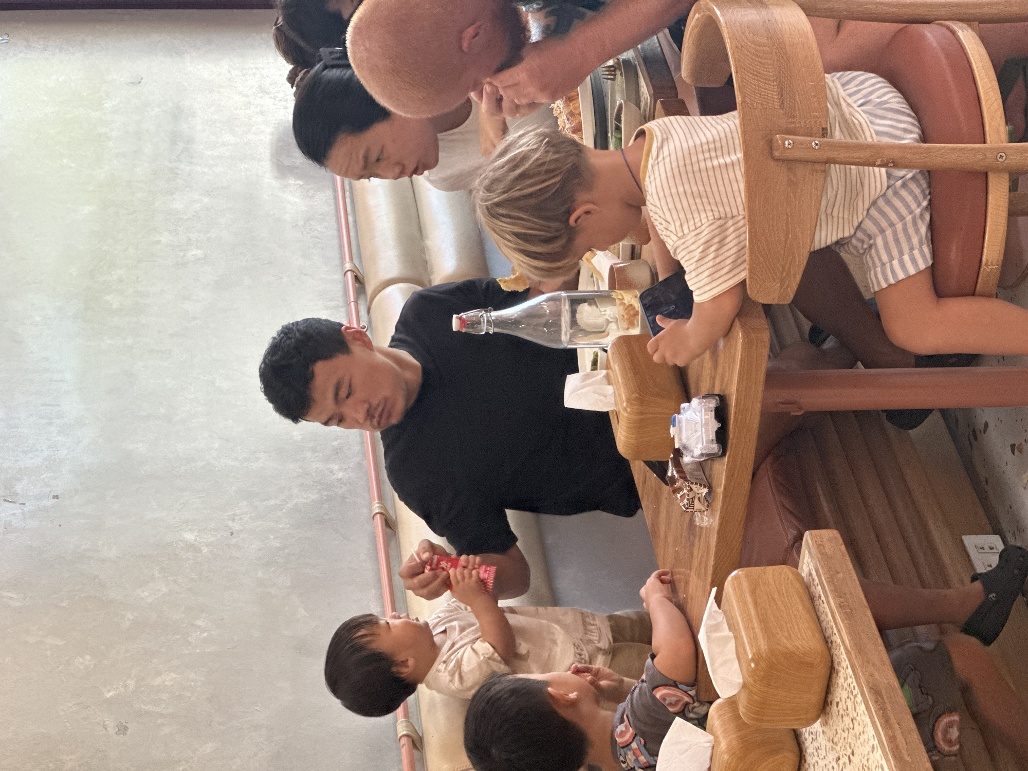 A group of people, including adults and young children, are seated around a wooden table in what appears to be an indoor restaurant with a textured concrete wall. In the foreground, a young child with blonde hair, wearing a striped t-shirt and blue and white striped shorts, is seated in a wooden chair and appears to be holding a dark object, possibly a phone. To their left, another child wearing a t-shirt with a superhero print is leaning on the table. An adult male in a black t-shirt is seated at the table, looking at a young child who is standing and holding a red object. To the right of the male, an adult female with dark hair is also seated at the table. Further to the right, another adult with blonde hair is partially visible. On the table are various objects including a bottle of water, food items, a tissue box, and toy cars. The overall setting suggests a casual family meal.