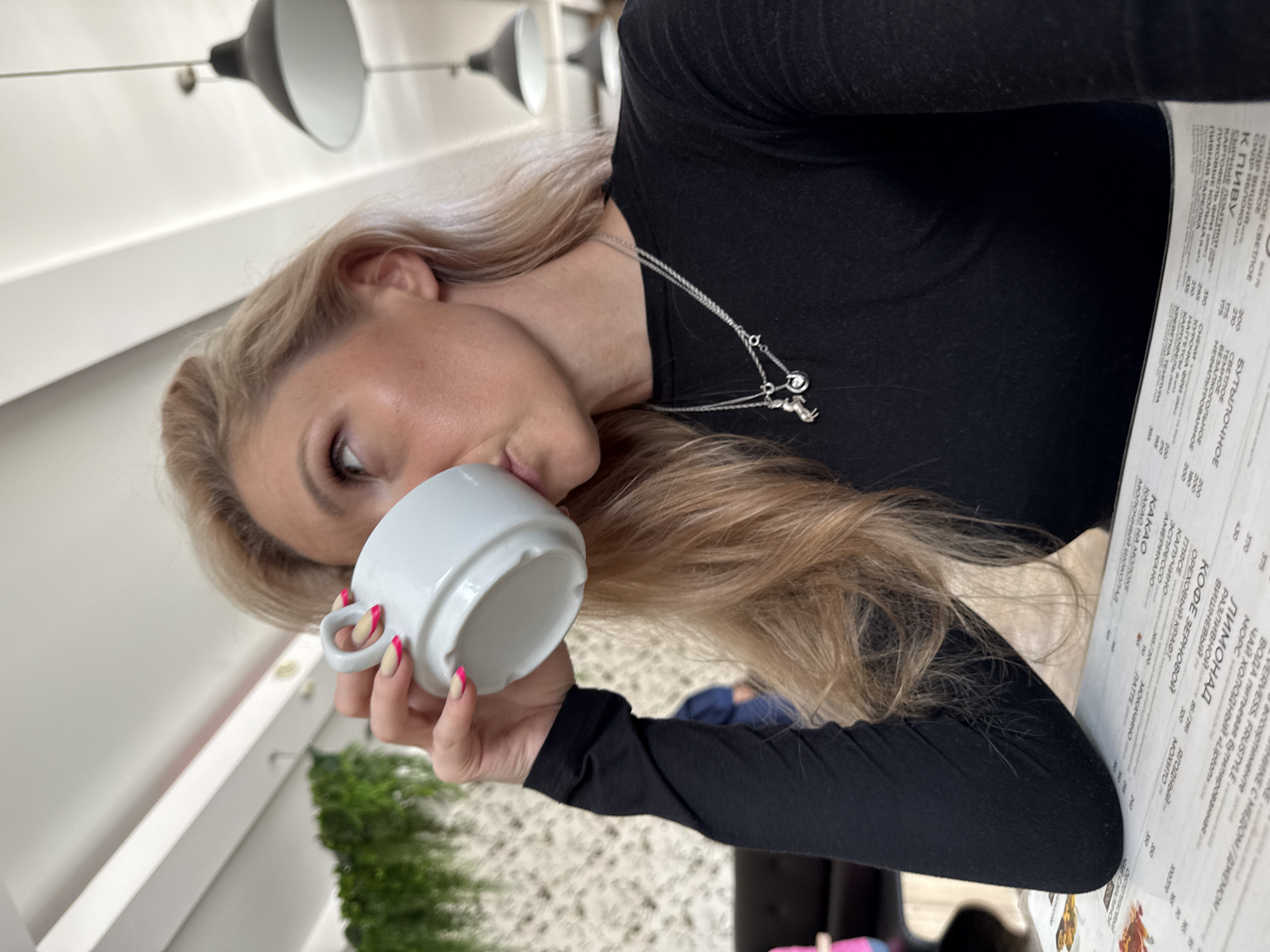 A blonde woman in a black long-sleeved top is drinking from a white mug. She is sitting at a table in what appears to be a cafe or restaurant, with a menu in front of her. The lighting suggests an indoor setting. The menu seems to be written in Russian. Through a window, there is some green vegetation. The woman's fingernails are painted with a pink accent. The location is likely Chaikovsky, Russia, based on the text of the menu.