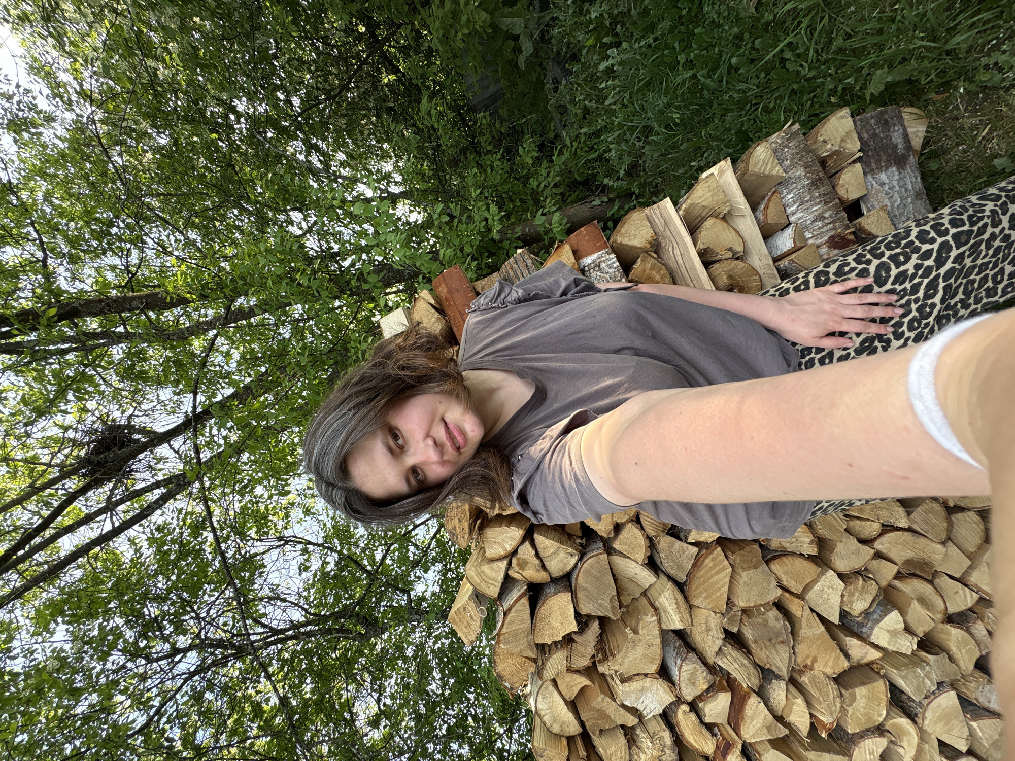 The image shows a woman lying on a pile of logs outdoors. The location is unspecified, and there is no information to suggest it is in Nizhnyaya Chermoda, Russia. The woman has brown hair with graying roots and is wearing a gray t-shirt and leopard-print pants. Her arm is extended towards the camera, suggesting she is taking a selfie. The background includes green foliage, possibly trees.