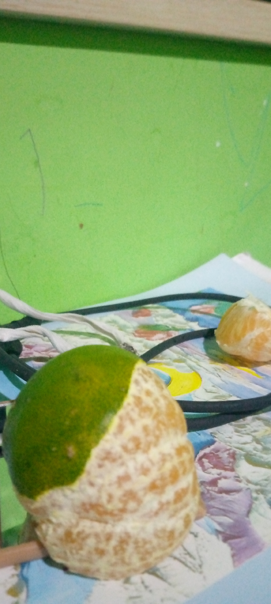 A close-up, low-angle shot shows a partially peeled orange against a light blue and white patterned surface. The orange is predominantly green on the left side, transitioning to an orange, peeled peel on the right. A thin wooden stick or skewer is visible protruding from the bottom left of the orange. In the background, a vibrant green wall with faint markings is visible. A black cord and a white cord are draped across the patterned surface, partially obscuring it. In the upper right corner, another section of peeled orange is visible. The scene is lit from above and to the right. The location is identified as Sumberwono, Indonesia.