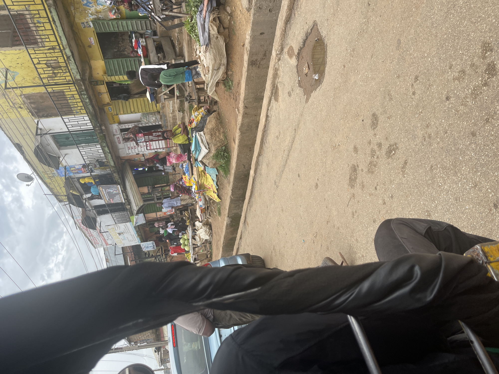 The image depicts a street scene in Gwong, Nigeria. In the foreground, a person is seated in what appears to be a tuk-tuk, with part of the vehicle's frame and the person's black clothing visible. The street is paved with asphalt and shows signs of wear, including a prominent pothole filled with muddy water. On the right side of the street, a row of buildings lines the sidewalk. These buildings have multiple stories with balconies, some with metal railings. The facades are painted in various colors, with a prominent yellow building dominating the scene. Shops are open on the ground floor, with green metal shutters. A market is set up along the sidewalk in front of the buildings. Various goods are displayed, including fruits like watermelons and bananas, as well as sacks of produce. Several individuals are present, some appearing to be vendors and others shoppers. A few people are standing or sitting near the market stalls. The sky is overcast.