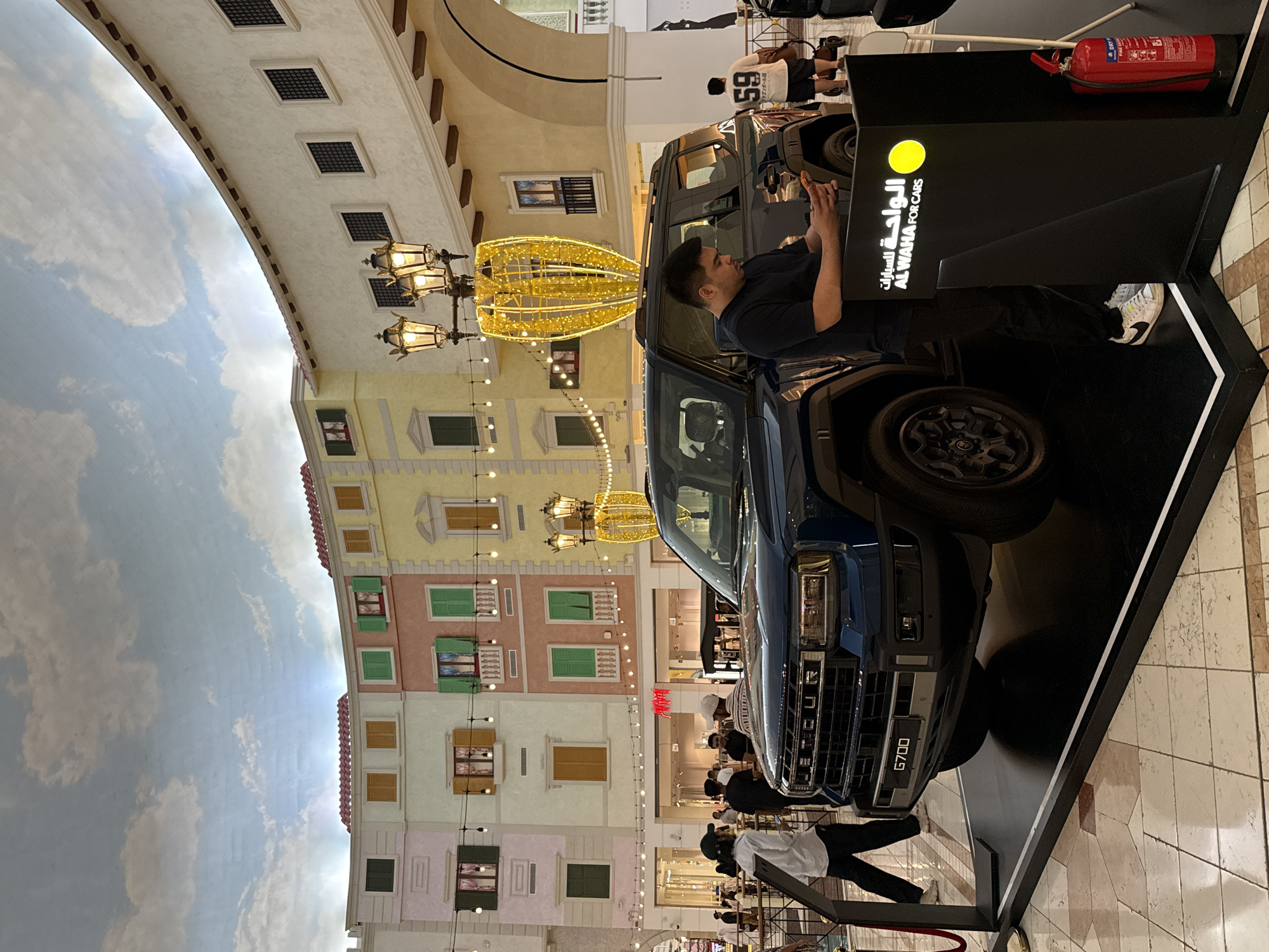 A man is examining a dark-blue SUV at the Villaggio Mall, in the city of Al Rayyan, Qatar. The SUV is displayed on a black platform with the dealership logo "AL WAHA FOR CARS". A fire extinguisher is also on the platform. There are many people shopping in the background. The mall is designed to mimic Venice, Italy, including buildings with painted facades and a painted sky on the ceiling.