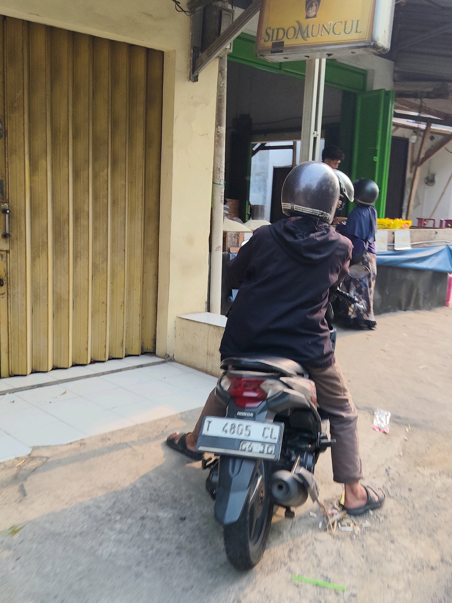 In Purwakarta, Indonesia, the image captures a street scene with two individuals on motorcycles. The foreground features a person from behind, wearing a dark hoodie and tan pants, seated on a black motorcycle. The motorcycle's license plate reads "T 4805 CL" and "06-20". The person is wearing a black helmet with a white stripe and dark-colored sandals. To the right of this motorcycle, another person in a blue shirt and patterned skirt stands near a blue tarp. In the background, a storefront with a green frame displays a yellow sign that says "SIDOMUNCUL". A yellow rolling door is visible on the left side of the image. The ground is a mix of concrete and pavement, with some debris scattered around. The lighting suggests it is late afternoon or early morning, with long shadows cast across the scene.