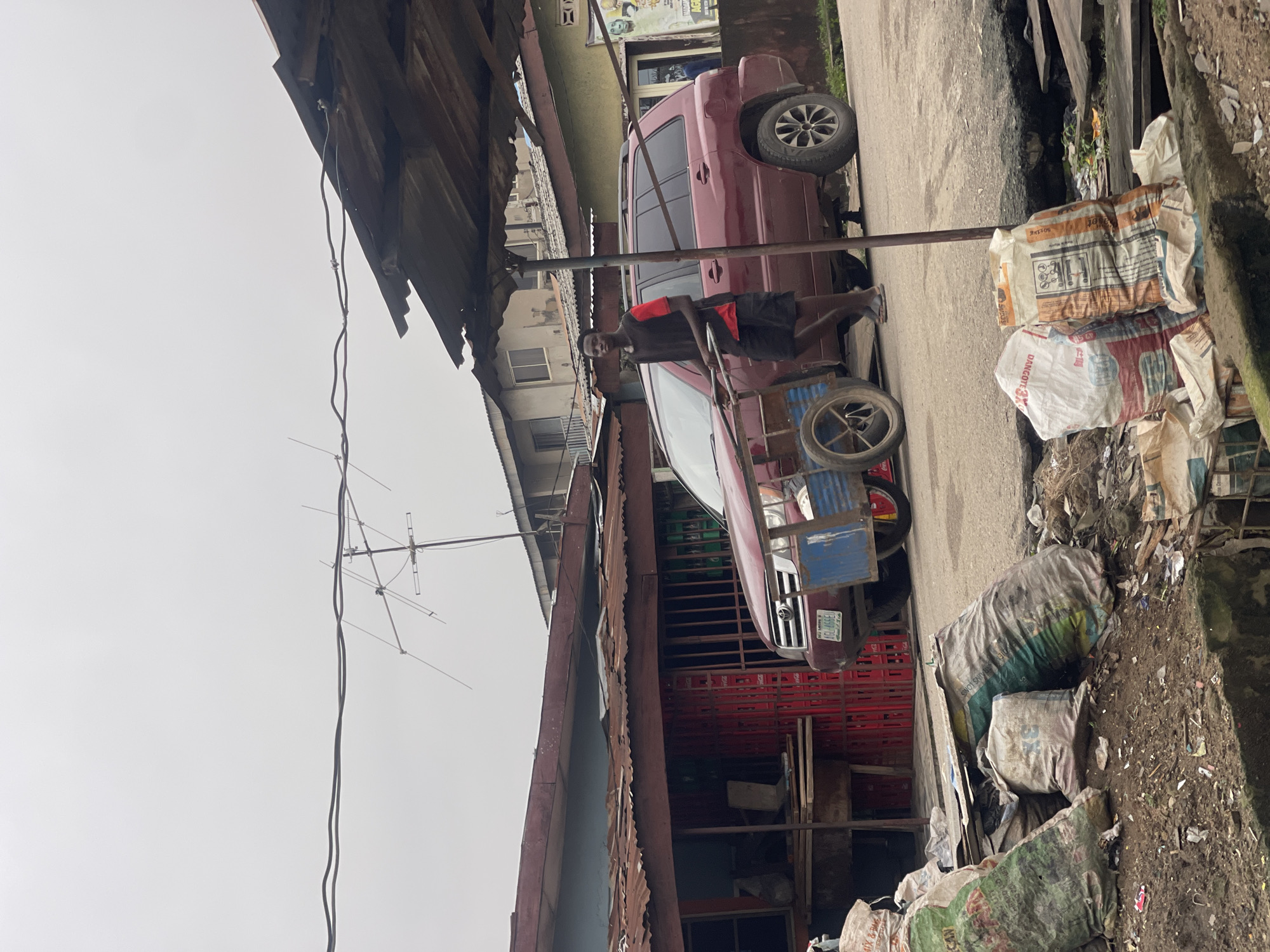 The image depicts a street scene in Akowonjo, Nigeria. In the foreground, several large sacks are piled on the ground, some appearing to contain debris or goods. A young man is walking in the middle ground, pushing a wheeled cart loaded with what looks like red crates. Behind the cart, a maroon SUV is parked. The man is wearing a black t-shirt with a red accent on the sleeve and black shorts. He is wearing flip-flops. The buildings in the background appear to be single or two-story structures with corrugated metal roofs. An antenna is visible on top of one of the buildings, and utility wires run across the top of the frame. The overall environment appears to be a bustling urban area with signs of commercial activity and some disarray, as suggested by the piles of sacks and the general condition of the buildings and road.