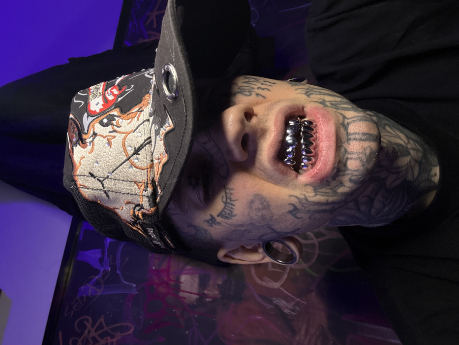 A person with extensive face tattoos wears a black baseball cap with detailed embroidery and metal grommets. The person has large gauges in their earlobes. Their upper and lower teeth are covered in chrome-like grillz. The background includes purple lighting and a reflective surface with abstract designs. The image is rotated.