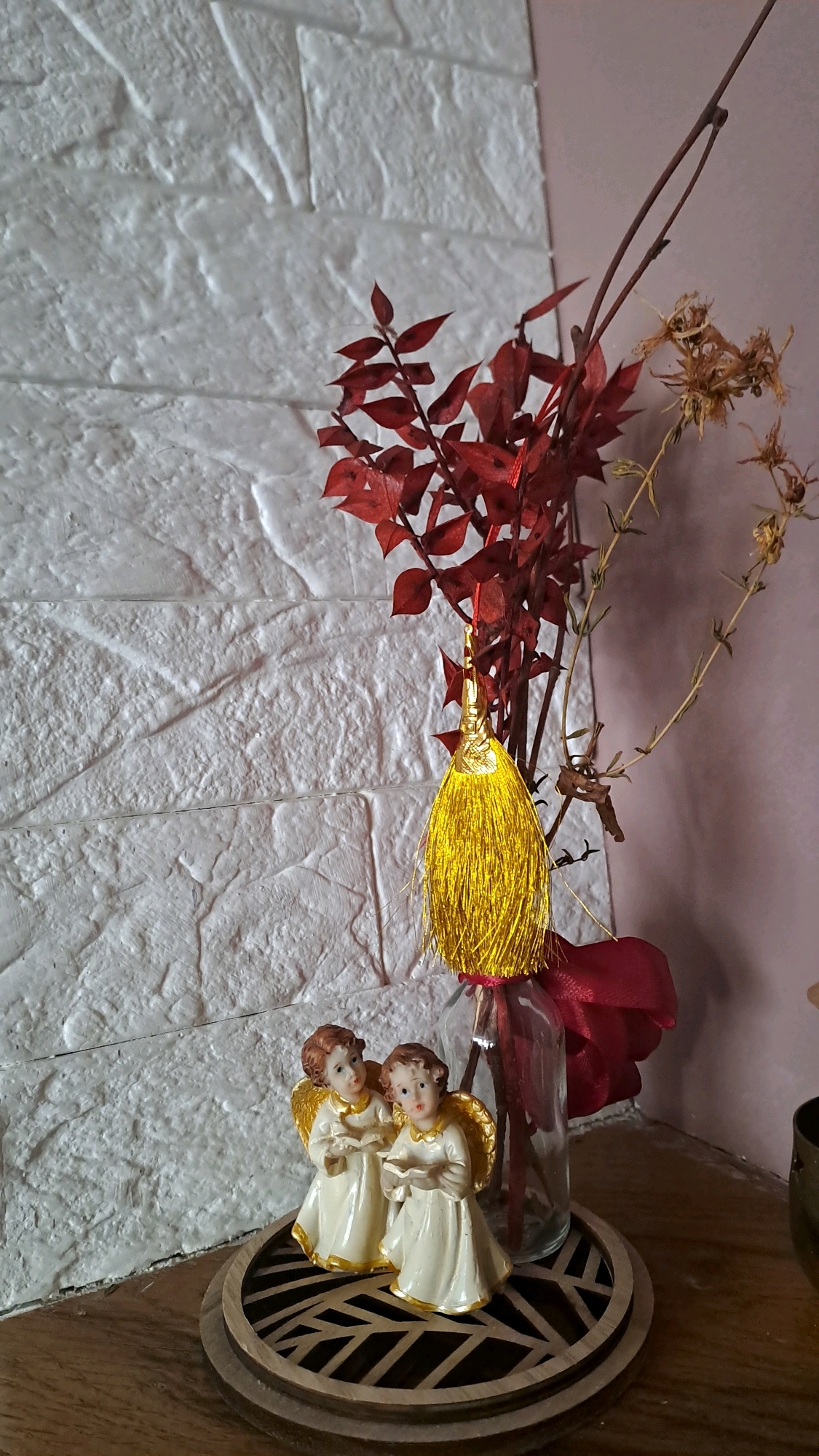 An indoor scene depicts two small figurines of cherubic angels with golden wings and halos, wearing cream-colored robes with gold trim. They are positioned on a round wooden coaster with an inlaid dark geometric pattern. Behind them, on the right side of the image, is a clear glass vase containing dried decorative branches with red leaves and some dried flowers. A large, golden tassel hangs from one of the branches. A red ribbon is tied around the neck of the vase. The background features a textured white wall with a brick-like pattern. The overall setting suggests a home interior, possibly a shelf or tabletop, with an autumnal or festive decoration. The photograph was taken in Kolomyia, Ukraine.