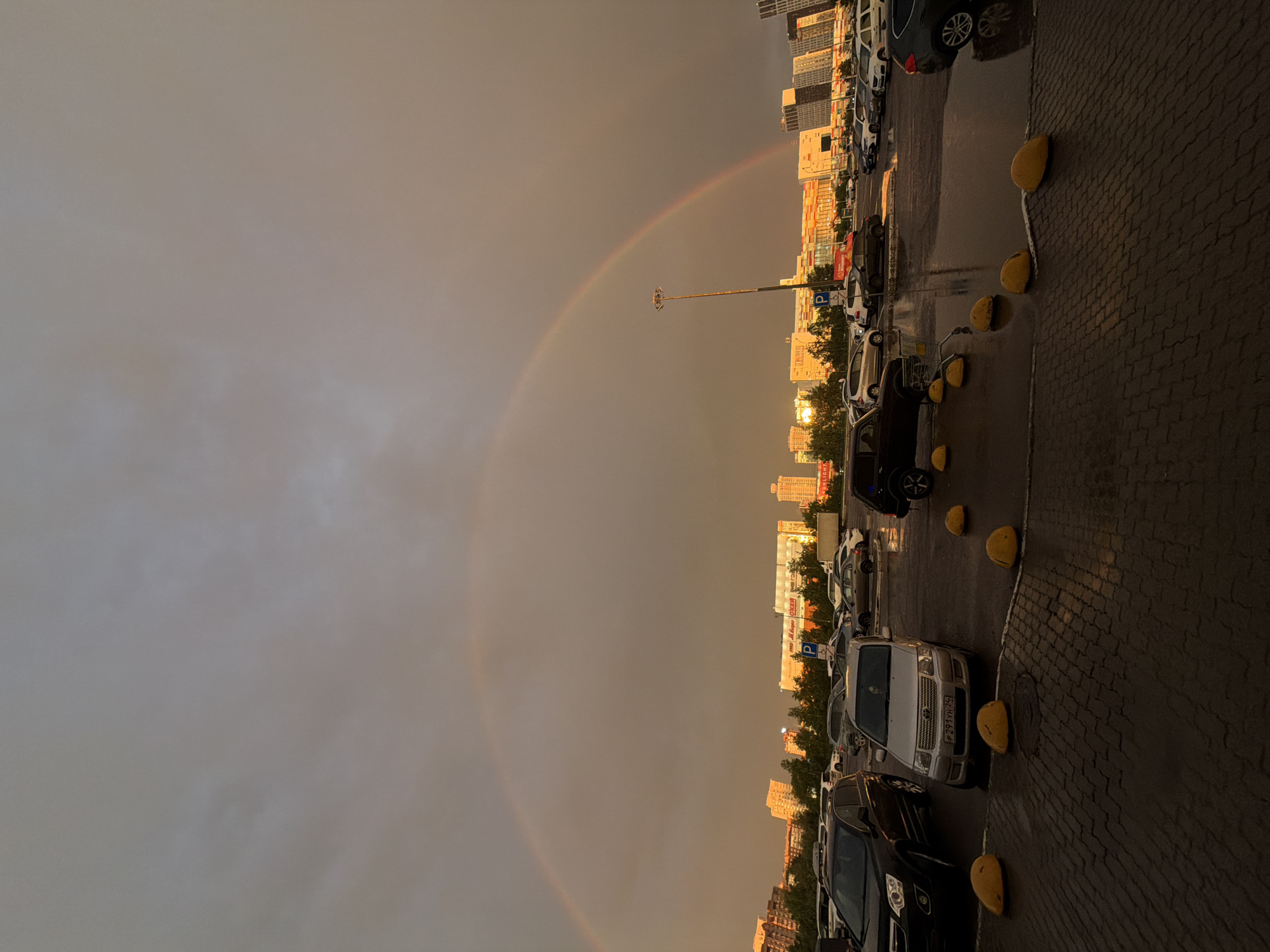 The image shows a parking lot in front of buildings in the city of Krasnoyarsk, Russia. A double rainbow arcs across the sky, with the sun setting behind a low-rise commercial building and apartment buildings. The parking lot is wet from rain, with puddles reflecting the sky and buildings. Several cars are parked in the lot, including a silver Toyota Probox in the foreground with the license plate "P 291YH 24". Yellow bollards are scattered throughout the parking lot. The sky is overcast and the light is dim, typical of a post-rain sunset.