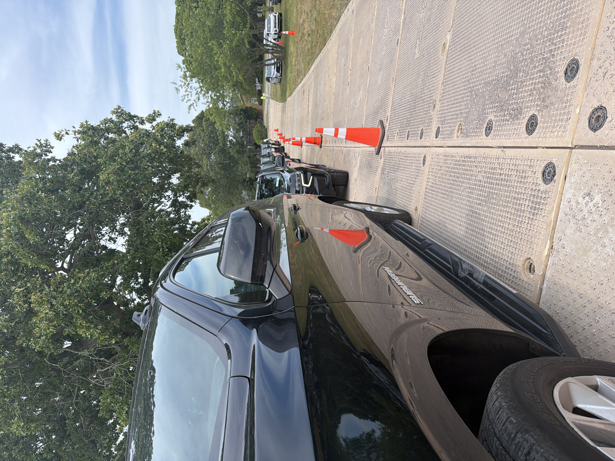 In the Town of Southampton, United States of America, a line of black SUVs is parked on a textured, tan surface, possibly a temporary roadway or staging area. Orange traffic cones are placed at intervals along the right side of the vehicles, indicating a defined pathway. The vehicles are facing away from the camera, towards a tree-lined area with a pale blue sky and scattered clouds overhead. The foreground shows a close-up of the front wheel and side of the closest black SUV, with the word "SUBURBAN" visible on its lower body. In the background, more vehicles and a few individuals are visible, with one person in a yellow vest standing near some white buildings. The overall scene suggests an organized event or a service being conducted outdoors.