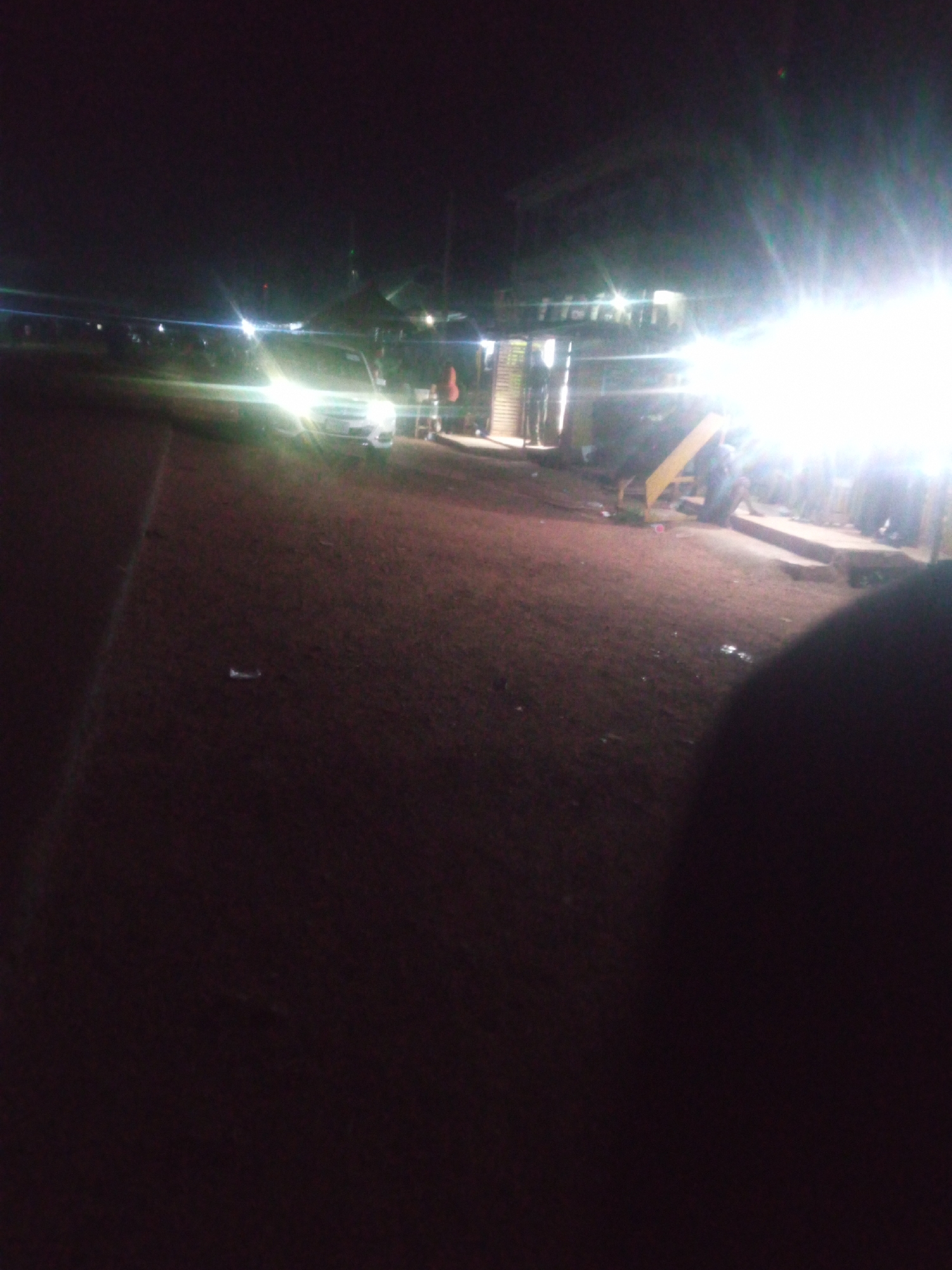 In an outdoor scene at night, an illuminated white van is parked on a dirt or gravel surface. The van's headlights are on, casting a bright glare. To the right of the van, a structure resembling a small building or stall with a sloped roof and a yellow ramp leading up to it is visible. Several individuals appear to be standing near the structure and on a raised platform. The overall lighting is dim, with strong light sources creating lens flares. The foreground is dark, with the camera operator's blurry shoulder visible on the right. The environment appears to be unpaved, possibly a marketplace or event space.