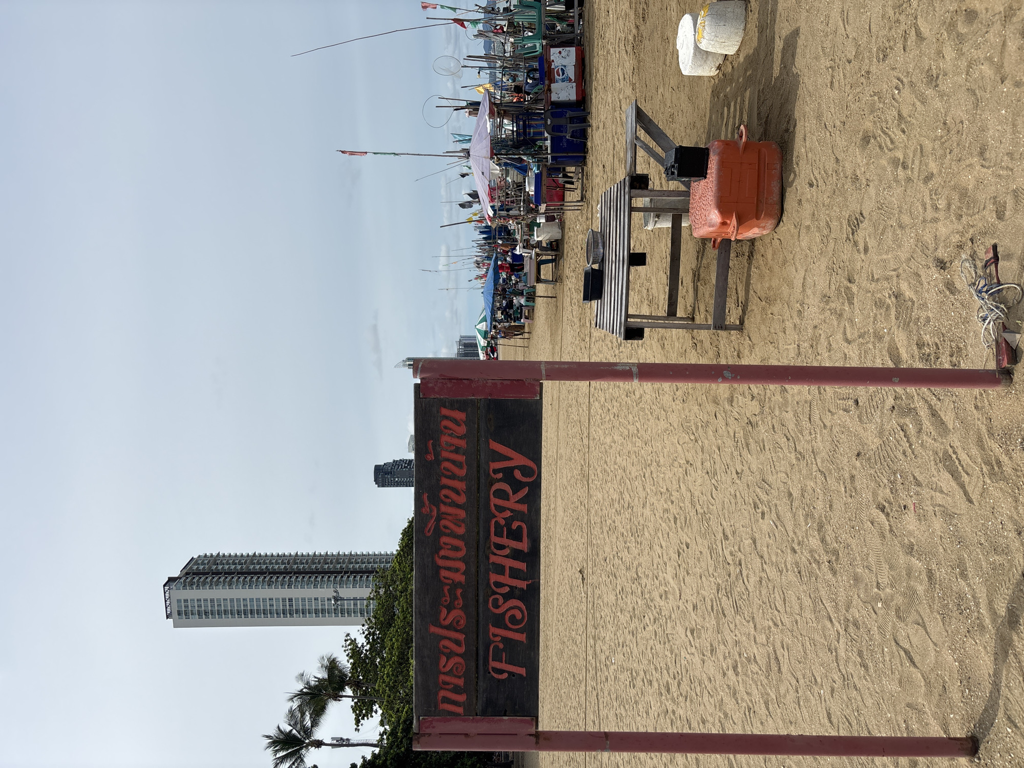 This image depicts a beach scene in Pattaya City, Thailand, with a focus on a wooden sign indicating "FISHERY" in English and Thai. The sign is mounted on red metal posts driven into the sand. In the background, a tall, modern skyscraper dominates the skyline, with palm trees and other greenery visible to its left. Further into the background, a row of stalls and vendors lines the beach, some with umbrellas and flags, suggesting a marketplace or activity area. Fishing equipment, nets, and boats are also visible amongst the stalls. In the foreground, close to the sign, is a simple wooden table with a few items on it, and an orange plastic container. The beach is sandy, with footprints indicating activity. The sky is overcast.