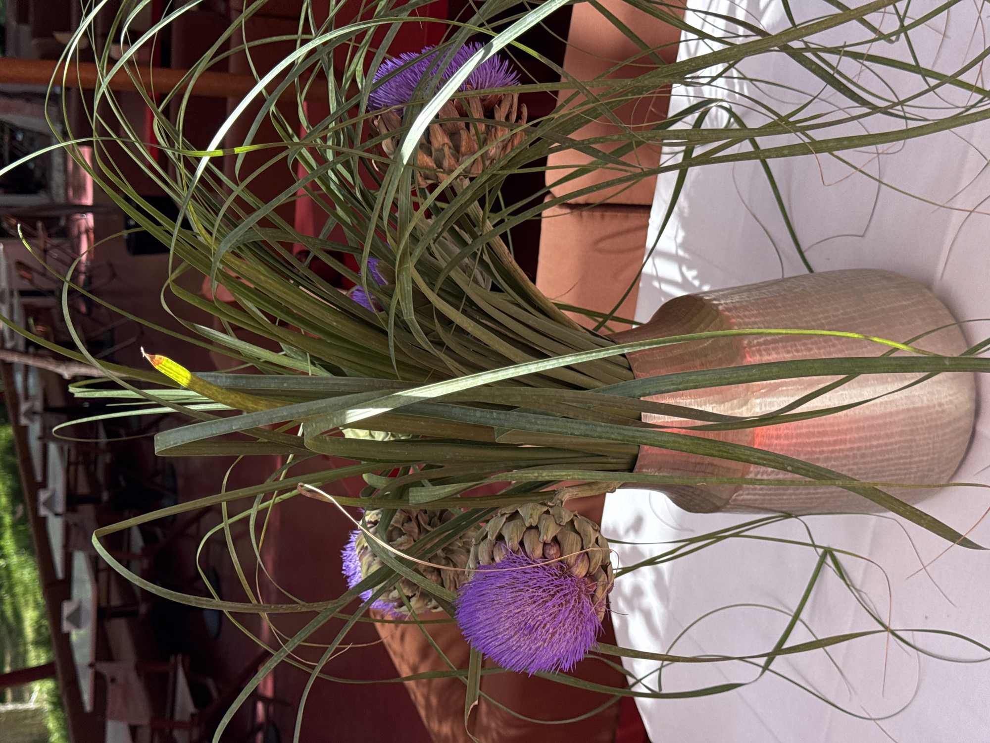 The image shows a floral arrangement in a metallic-toned vase. The arrangement features spiky green foliage, as well as what appear to be purple thistle-like flowers. The vase is placed on a white tablecloth. The background suggests an outdoor or semi-outdoor setting, possibly a restaurant or event space, with tables and chairs visible to the left. The lighting indicates it is daytime. There are no people visible. The location is Yerevan, Armenia.