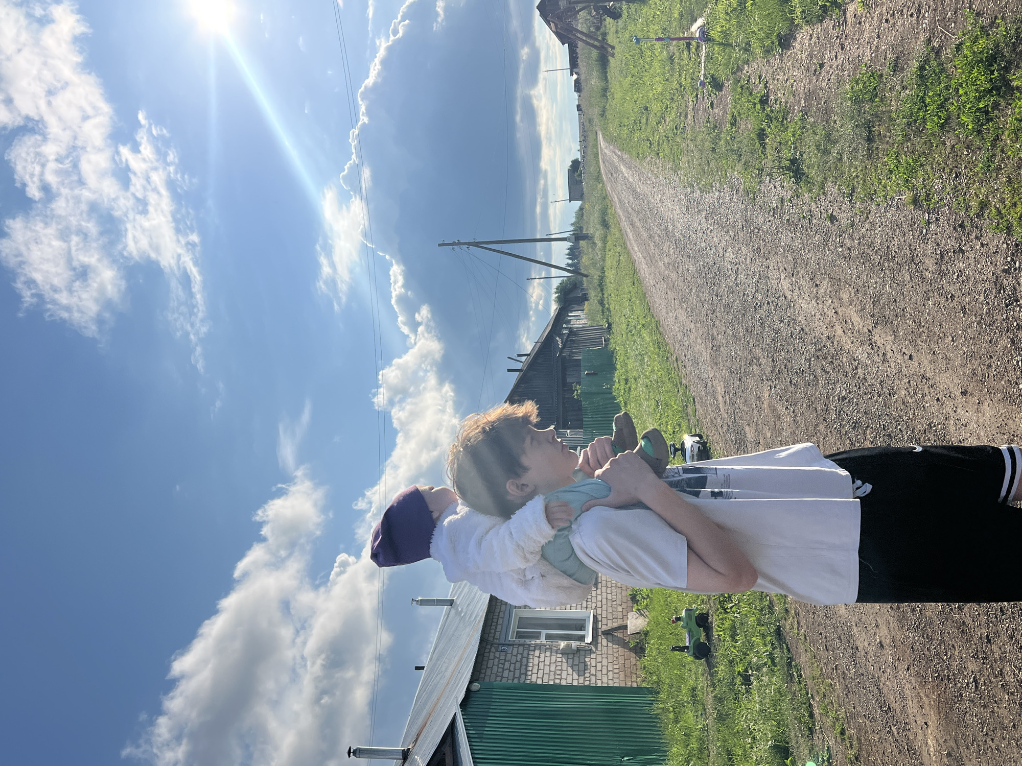 In Panteleevka, Russia, a young boy carries a baby on his shoulders down a gravel road. The boy wears a light-colored t-shirt and black shorts. The baby is dressed in a white jacket and a purple hat. They are positioned in the foreground, facing away from the camera, towards a rural landscape. The environment features a dirt road stretching into the distance, flanked by green grass and scattered wooden houses. A large, fluffy cloud dominates the blue sky, with the sun shining brightly, casting a lens flare across the upper right quadrant of the image. The scene suggests a warm, sunny day in a village setting.