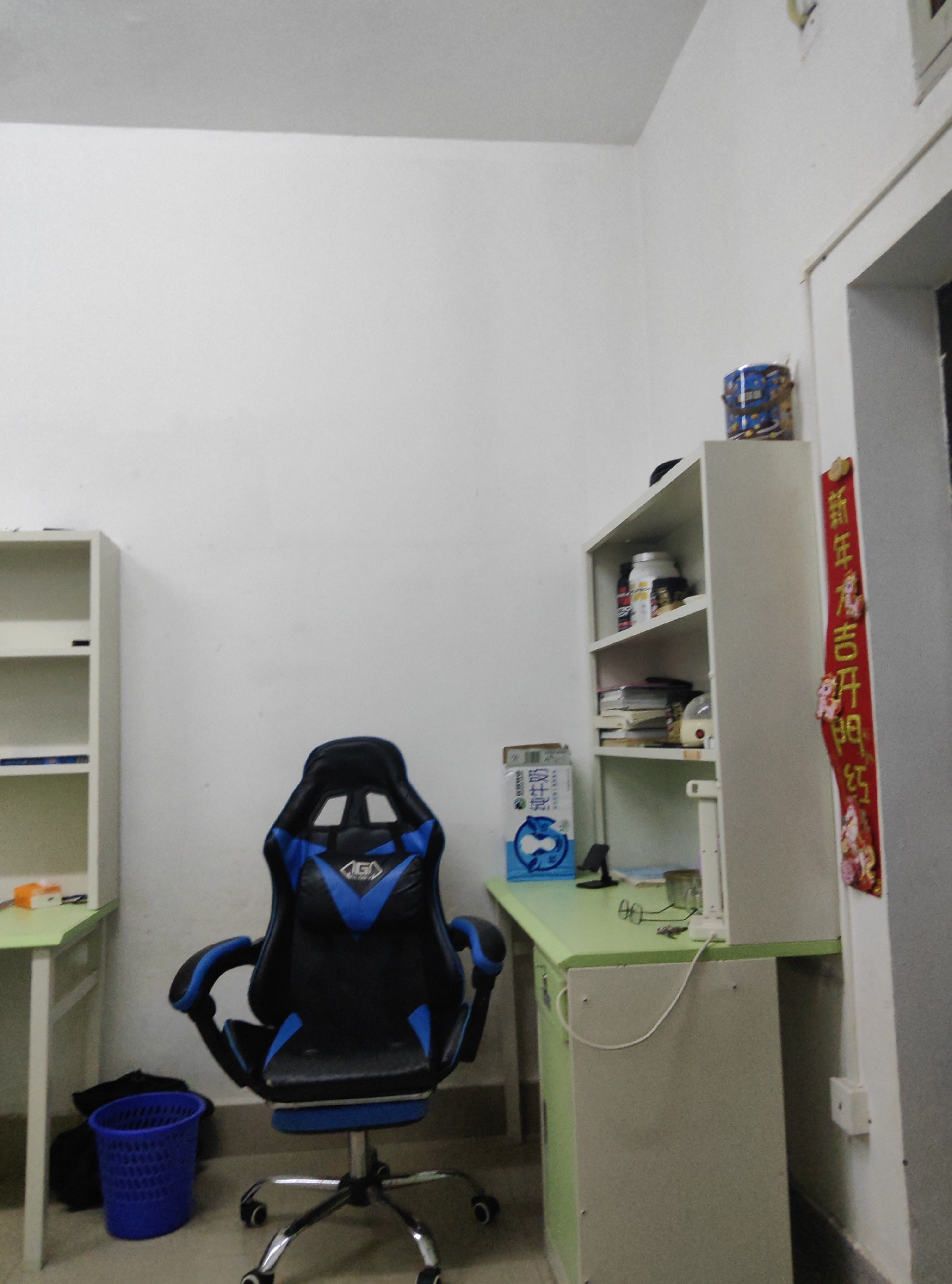 This is a photograph of a room, likely a student's dorm room or small office, located in the Chongqing High-tech Zone, China. The room is sparsely furnished and appears to be tidy.
The primary focus of the image is a black and blue gaming chair, centrally positioned in the room. The chair is on casters and has a distinctive ergonomic design. To the left of the chair is a white bookshelf with several shelves. The shelves contain various items, including what appear to be books, containers, and possibly some decorative items.
To the right of the gaming chair is a green desk with a filing cabinet underneath. On the desk, there are a few items, including what looks like a pair of glasses and a small blue fan. A red banner with Chinese calligraphy hangs vertically on the wall to the right of the desk.
In the lower left corner of the image, a blue plastic trash can is visible. A black bag is placed next to the trash can. The walls of the room are painted white, and the floor is a neutral color. The lighting in the room is bright, suggesting it is daytime or the room is well-lit by artificial lights.
The overall impression of the room is functional and uncluttered, with a focus on study or work. The presence of the gaming chair suggests the occupant may be a student or someone who enjoys gaming. The room's location in the Chongqing High-tech Zone implies it is likely within a modern urban environment, possibly a university campus or residential area catering to professionals.