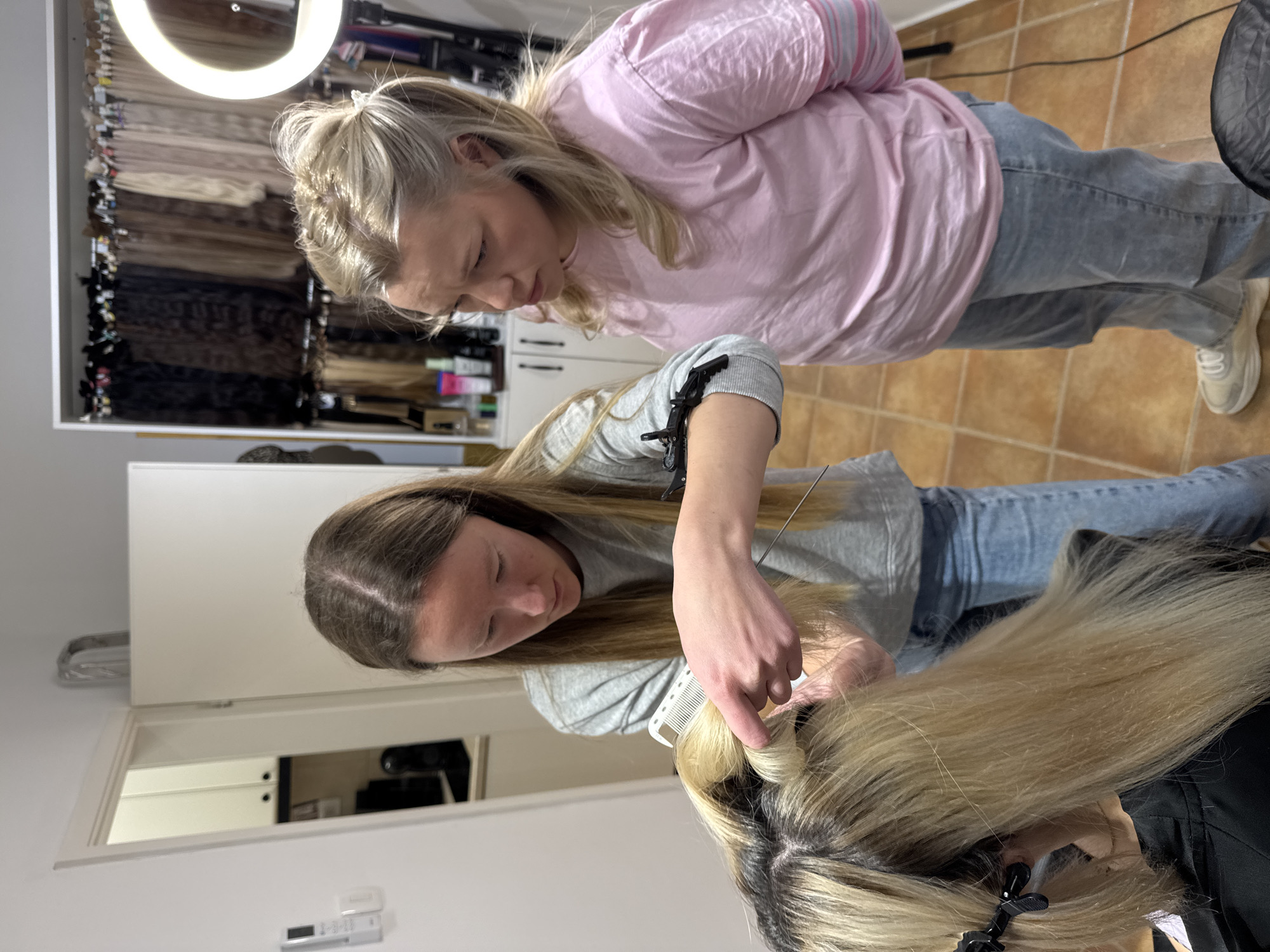 Two women are indoors, likely at a hair salon. One woman, wearing a light gray t-shirt and jeans, is carefully sectioning and combing through a client's blonde hair. The client is seated, and the back of her head and hair are visible, with clips securing sections. The second woman, wearing a pink long-sleeved shirt and jeans, stands behind the first, observing or assisting. In the background, there's a display of what appears to be hair extensions of varying shades, and a ring light. The location appears to be a professional or well-equipped home salon.