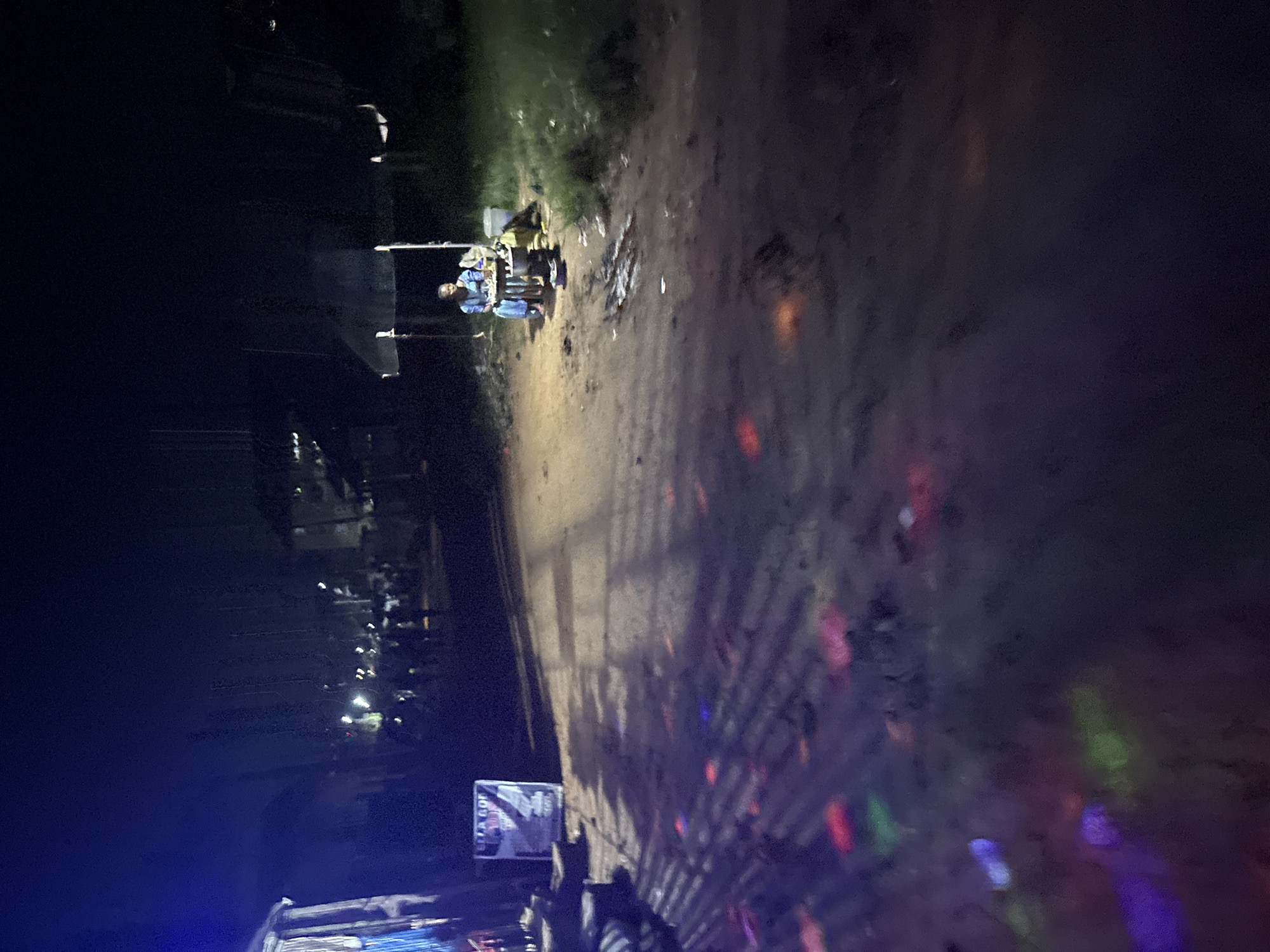 The image depicts a nighttime street scene in Nkpor, Nigeria. A dirt road is visible, illuminated by colored lights projected onto its surface, suggesting a festive or event atmosphere. To the right, a person is seated at a small table under a canopy, with a pot or stove in front of them. Behind them, a bucket and other items are present. Further down the road, more people are faintly visible in the distance, along with structures that appear to be buildings or stalls. A sign is posted on the left side of the road. The overall impression is that of a lively local gathering or market at night.