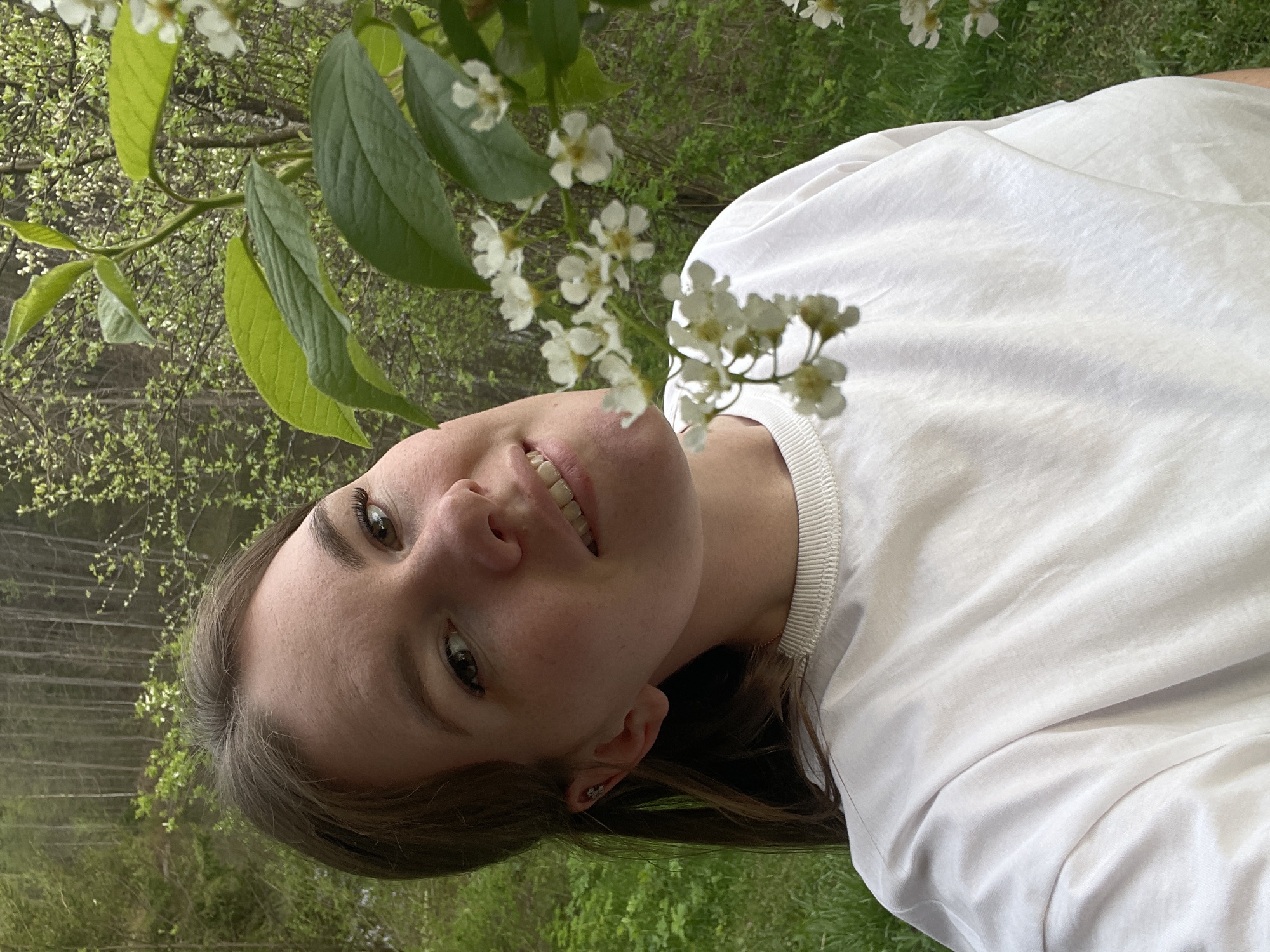 A young woman is posing outdoors with branches of white flowers and green leaves framing her face. She is wearing a white t-shirt. The background appears to be a wooded or grassy area. The location information is not present in the image itself.