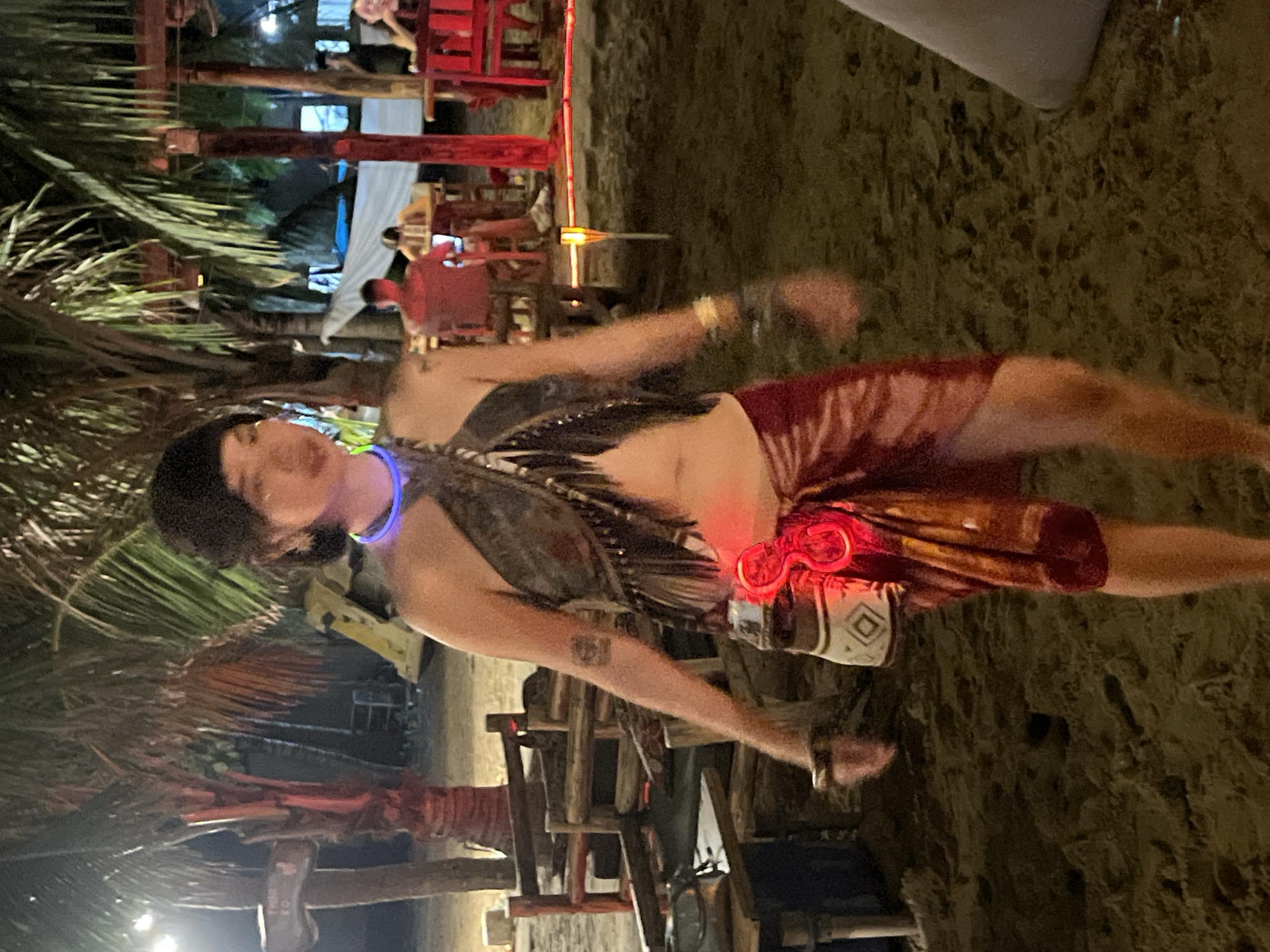 The image shows a person performing on a beach at night, likely in Đà Nẵng, Vietnam. The person is wearing a headdress and a costume made of feathers, a chest plate, and tribal-inspired shorts. They are also wearing a blue neon necklace and have a red light-up accessory around their waist. The person appears to be dancing or performing, with their arms extended. The background shows a beach setting with palm trees, sand, and wooden structures that resemble tables and chairs, suggesting a beachside venue. Red lights illuminate some of the background structures. The time of day is clearly night, with the scene illuminated by artificial lights. The overall atmosphere appears festive and energetic, possibly indicating a cultural performance or a night market event.