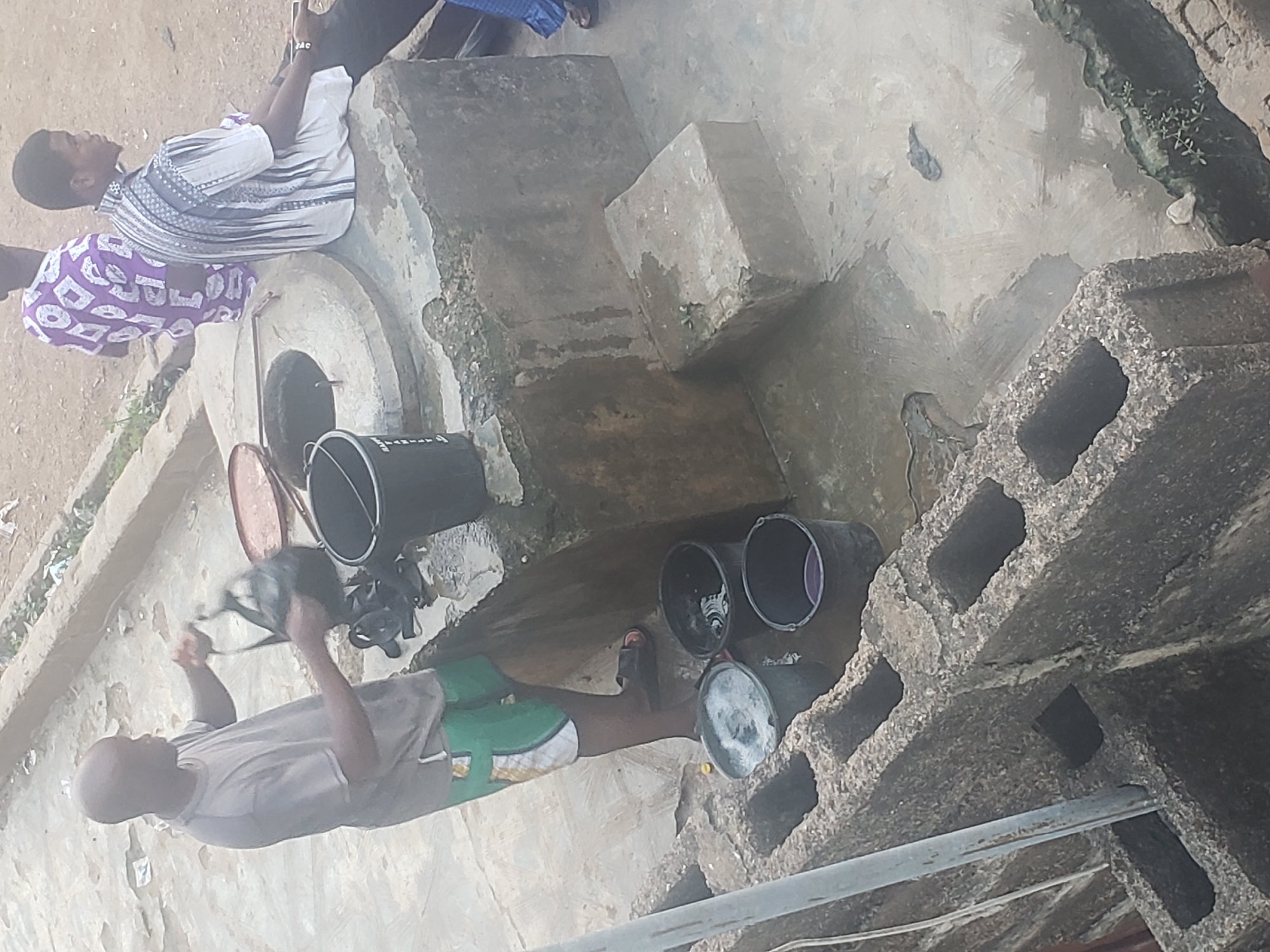 In Alalubosa, Nigeria, a high-angle shot depicts a concrete structure, likely a well, where multiple black buckets are positioned. One man is actively drawing water from the well using a bucket, while two other men are standing nearby, observing. The surrounding environment appears to be a residential area with basic infrastructure.