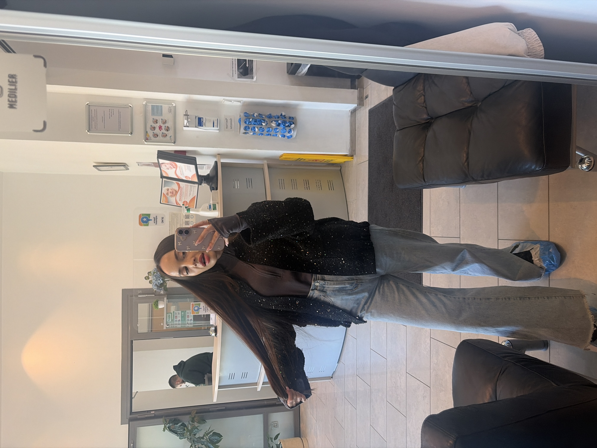A woman stands in the hallway of a MEDILIER-branded medical clinic, taking a selfie in a mirror. She is wearing a brown top, jeans, and a sparkly black jacket. Blue shoe covers are visible on her feet. In the reflection, another person is visible behind a counter. The environment includes a check-in desk, seating area with black leather chairs, and informational posters on the wall. The clinic appears to be clean and well-lit. Based on the branding, this photo may be taken at a MEDILIER location in Saint Petersburg, Russia.