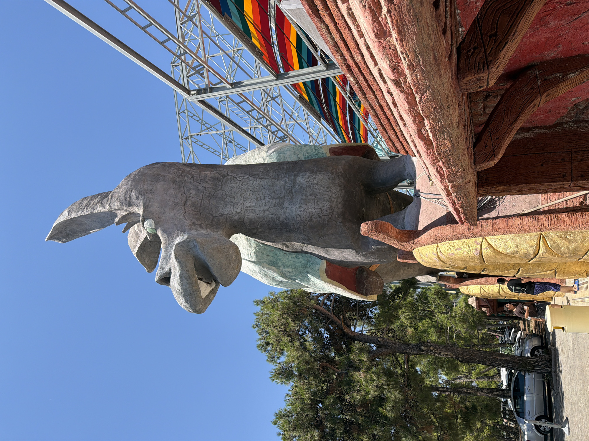 An outdoor, low-angle, full shot captures a large, gray, dinosaur-like sculpture with a light green and red chest, standing in front of a bright blue sky. The sculpture's mouth is open, revealing white teeth. Behind the sculpture, a metal framework supports a colorful, striped canopy. To the left, a cluster of green pine trees stands next to a parking lot with several cars. In the foreground, a portion of a red, wooden-like structure with a textured roof is visible. Two young men are standing near the base of the sculpture, with one holding a drink and the other looking towards it. The scene is located in Kemer, Turkey.