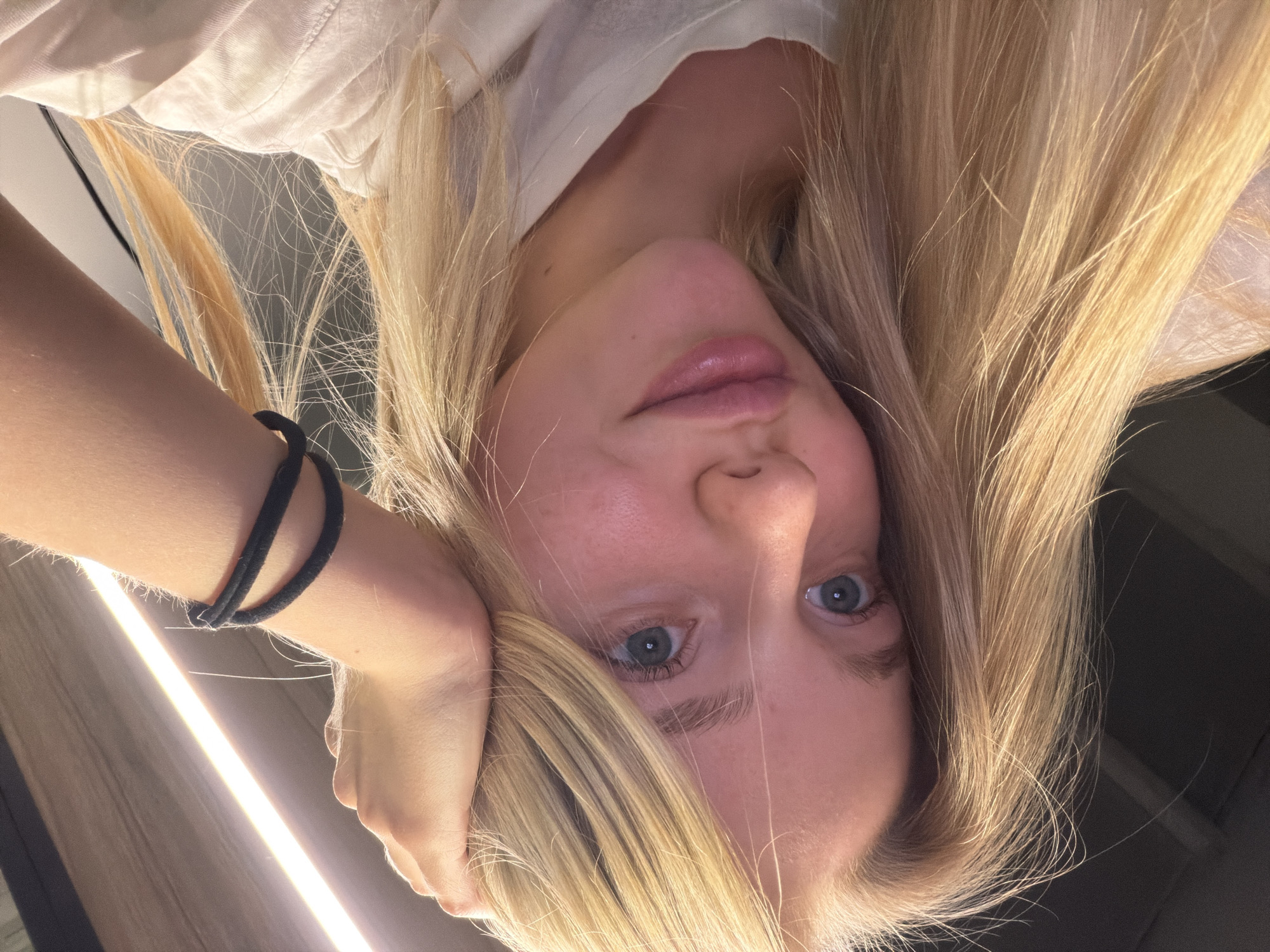 An overhead shot looking down at a young person with long, blonde hair. The person is fair-skinned with blue eyes and light pink lips. Their arm is raised, with their wrist bent and hand touching the side of their face. A black hair tie is around their wrist. The person is wearing a light-colored top. The background appears to be an indoor setting with a light fixture. The location is Unknown, Unknown.
