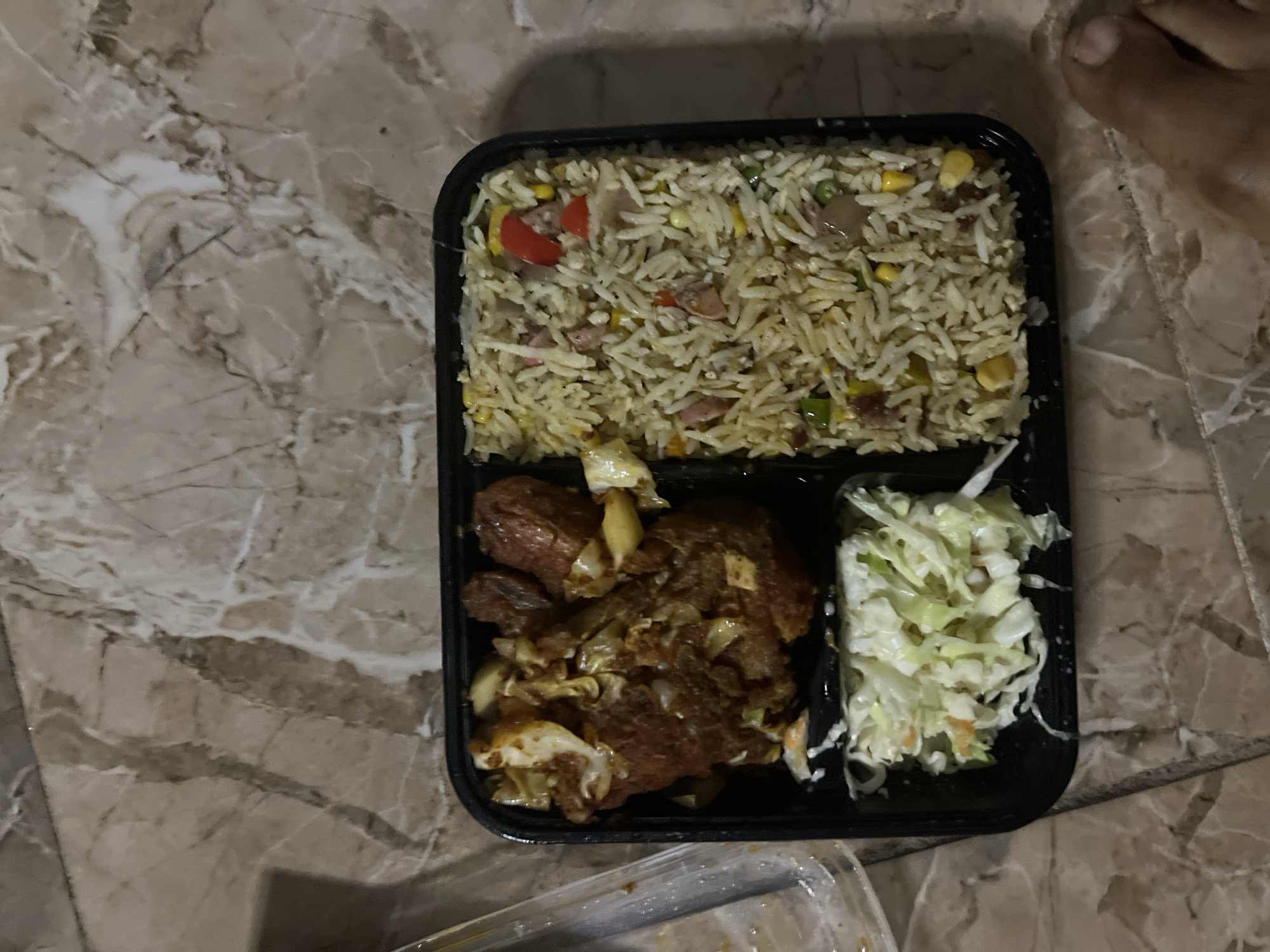 A top-down, close-up shot reveals a black, three-compartment food container. The container is placed on a marble-patterned surface. The largest compartment contains a pile of fried rice with visible specks of corn, peas, and red peppers. Another compartment holds pieces of fried meat mixed with what appears to be sautéed onions. The smallest compartment contains coleslaw. A portion of a human foot is visible in the upper right corner of the image. The photograph was likely taken indoors in Jalingo, Nigeria.