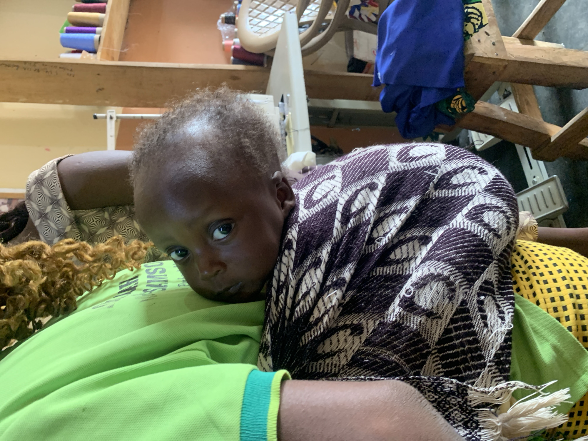 The image depicts a close-up view of a baby being held by a person. The baby, positioned in the foreground, has dark skin and is looking towards the camera with wide eyes. The baby is wrapped in a patterned cloth, with fringes visible at the bottom. The person holding the baby is wearing a bright green shirt with a striped collar. The background suggests an indoor setting, possibly a workshop or a room with sewing materials. A sewing machine is partially visible, along with spools of thread on a shelf and a wooden stool with fabric. The overall lighting is natural, casting soft shadows. The image appears to be taken in Jos, Nigeria, though no specific landmarks confirm this.
