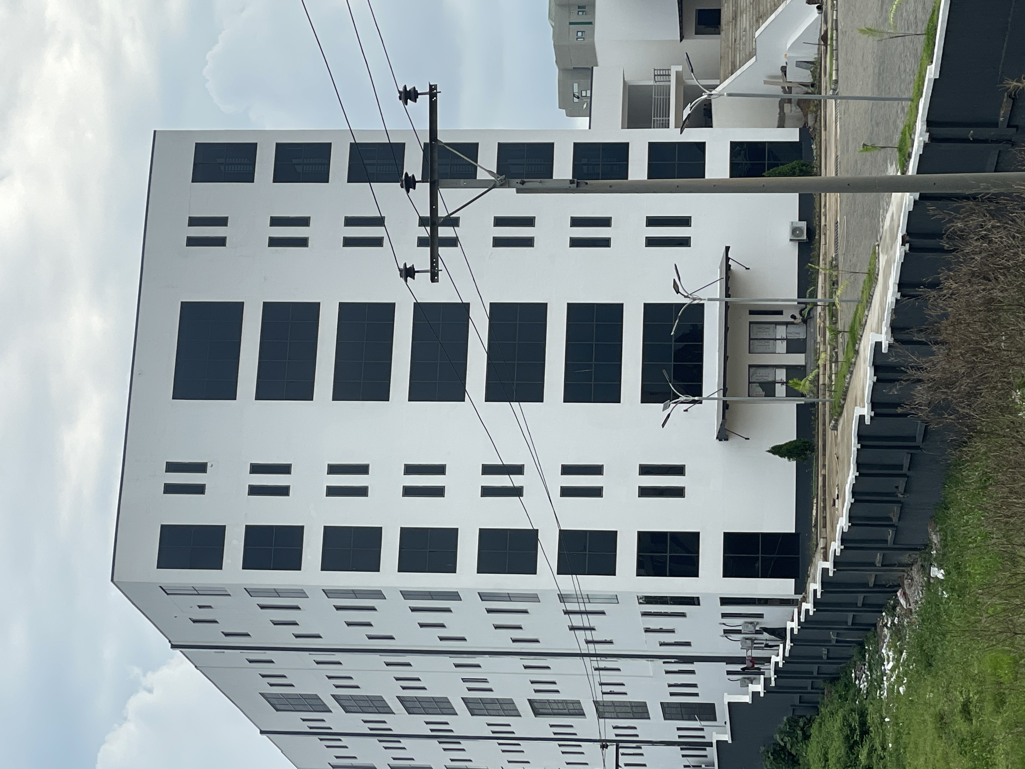 The image captures a modern, multi-story white building in Abuja, Nigeria, characterized by its minimalist design and numerous dark, rectangular windows. The building has a stark, geometric facade with rows of large square windows and smaller, vertical rectangular windows. The overall impression is one of contemporary architecture. In the foreground, a dark gray fence with white accents runs horizontally across the image, separating a patch of overgrown green grass and scattered debris from a paved area in front of the building. Beyond the main building, another section of the structure is visible, featuring a white staircase leading to an upper level. Power lines with ceramic insulators run diagonally across the sky, supported by a utility pole situated in front of the building. The sky is overcast with scattered clouds, suggesting a cloudy day. A few small trees and streetlights are also present around the paved area. Two individuals are faintly visible near the entrance of the building, one seated and another standing. The scene appears to be a commercial or office complex in a developing urban area.