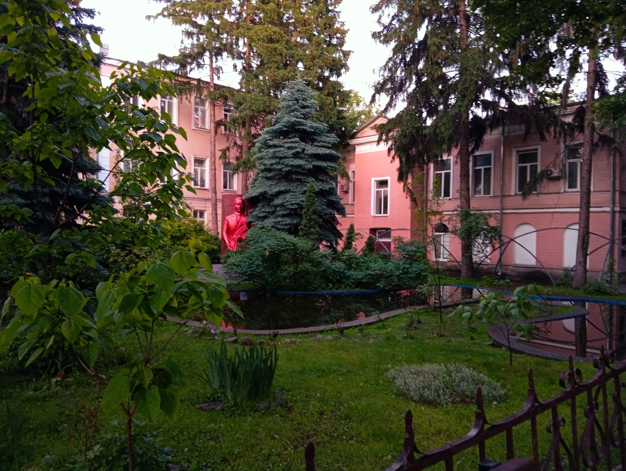 The image depicts a garden in front of a pink building, likely located in Poltava, Ukraine. The garden features a variety of trees, bushes, and flowers. A bright red statue is prominently displayed amidst the greenery. A dark wrought-iron fence runs along the front edge of the garden. The pink building behind the garden has multiple windows and a distinctive architectural style.