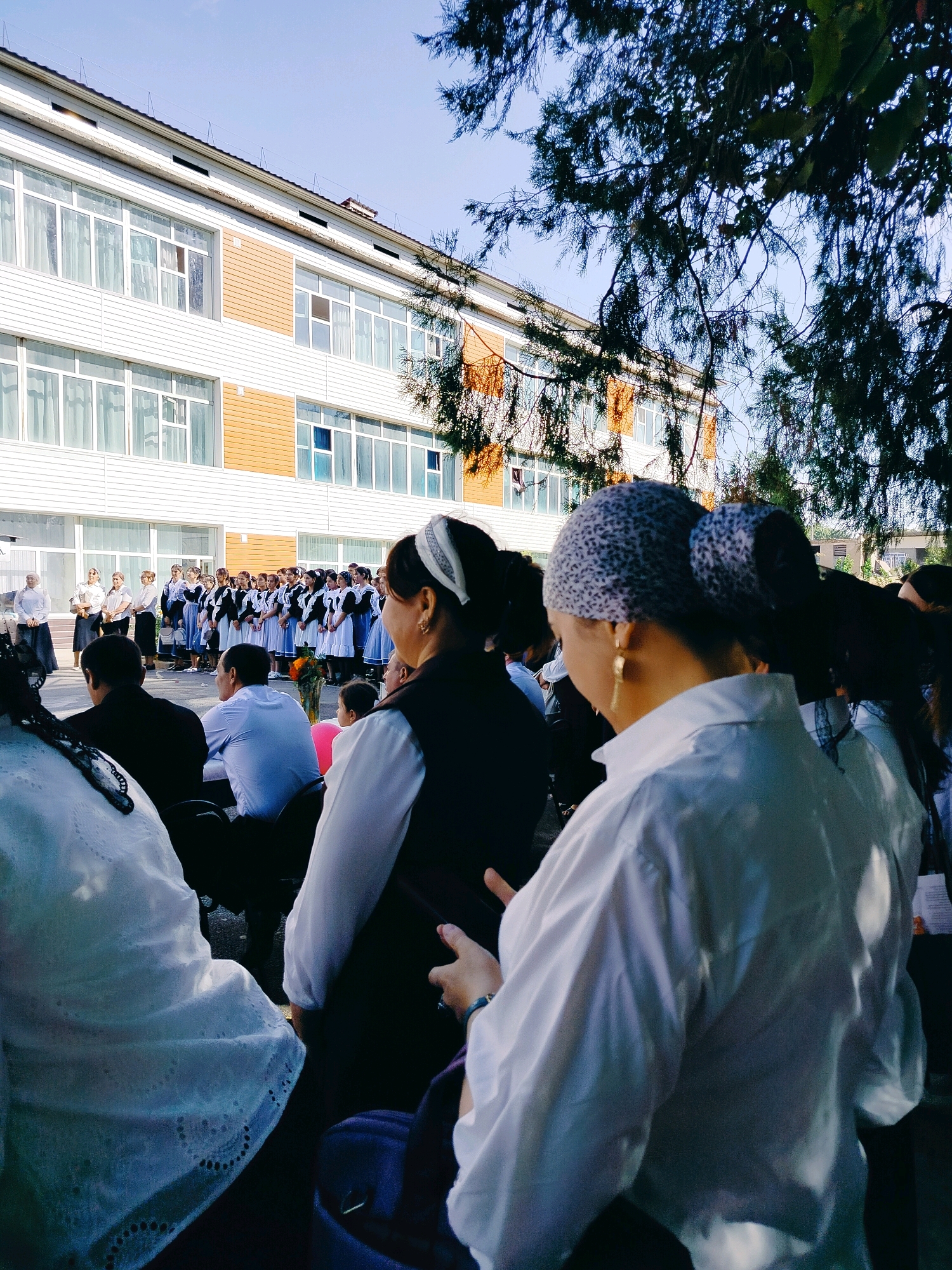 The image depicts an outdoor scene in front of a modern school building with white and orange siding and large windows, located in the city of Kölkent, Kazakhstan. In the foreground, several people are visible from behind, with a focus on two women wearing headscarves and casual attire. One woman on the right has a grey and black patterned headscarf and large gold hoop earrings, and is wearing a white collared shirt. Another woman on the left is wearing a white lace top and a black headscarf. Behind them, a group of young people, likely students, are standing in formation. They appear to be dressed in school uniforms, with many wearing black vests over white shirts and dark skirts or pants, and some wearing white headscarves. Adults, possibly teachers or parents, are also present, some standing and some seated. The environment suggests an event or gathering taking place outside the school, possibly a ceremony or assembly, under a bright sky with tree branches visible in the upper right corner.