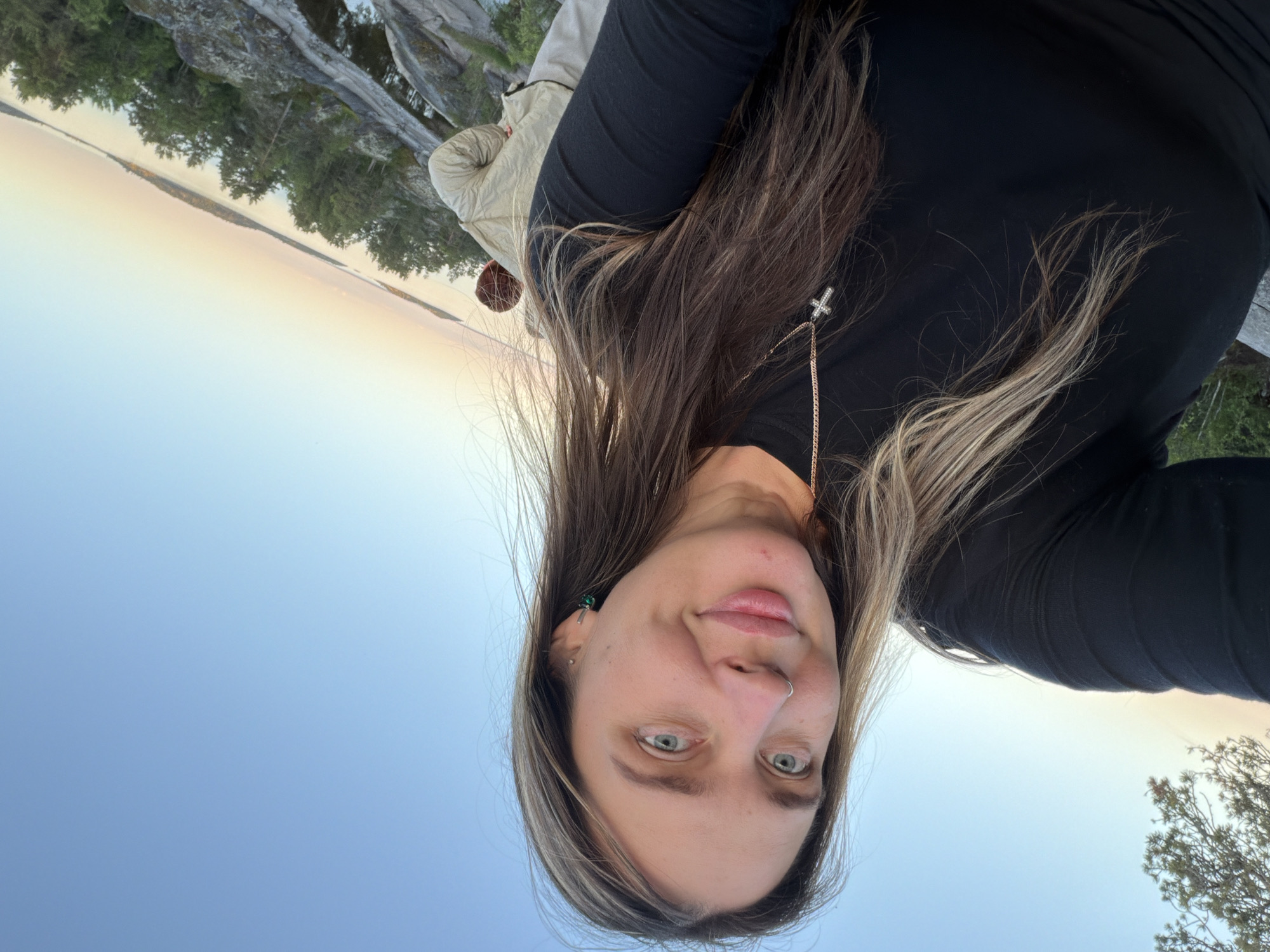 A medium shot selfie shows a woman with light brown hair and blue eyes posing outdoors. She is wearing a black long-sleeved shirt and a necklace with a cross pendant. She has a nose ring and an earring in her left ear. Behind her, a body of water can be seen with islands covered in trees. The sky is clear and blue, with a hint of orange from a sunset. Another person, wearing a light-colored jacket, is visible in the background. The location appears to be a rocky area with trees, possibly on a hill overlooking the water.