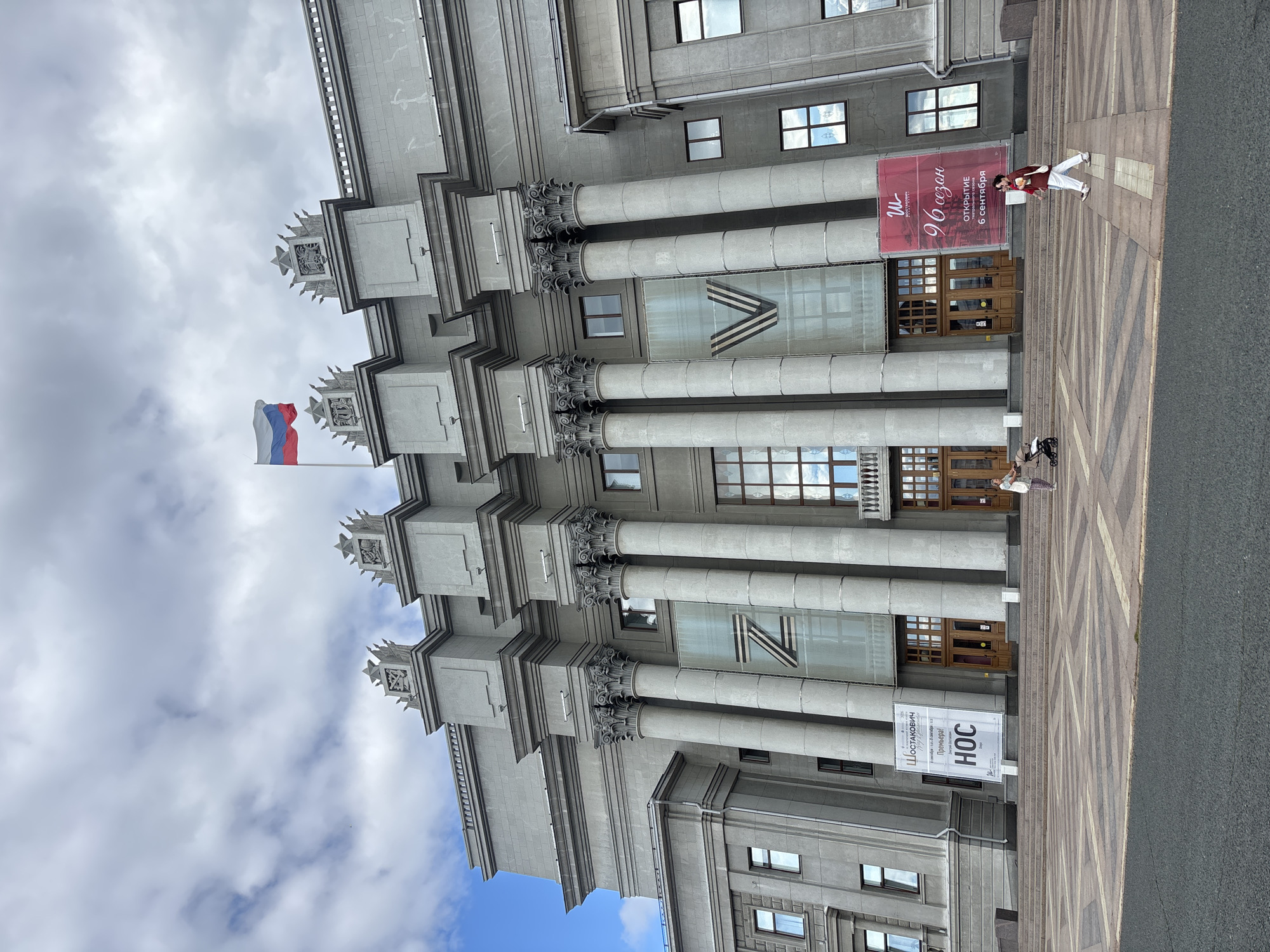 The image shows a large, imposing grey building with classical architectural elements, including large columns and decorative stonework. A Russian flag flies from a pole on the rooftop. Two large banners are displayed on the facade, one with the letter "Z" and another with the letter "V". In front of the building, there is a wide paved plaza with geometric patterns. Several people are visible on the plaza. On the left, a person pushes a stroller. On the right, a person walks past carrying a bag. Two smaller banners are placed at the base of the steps leading up to the building, advertising an event or performance. The sky above is partly cloudy. This building is the Novokuybyshevsk Oil Refinery Palace of Culture in Novokuybyshevsk, Russia.