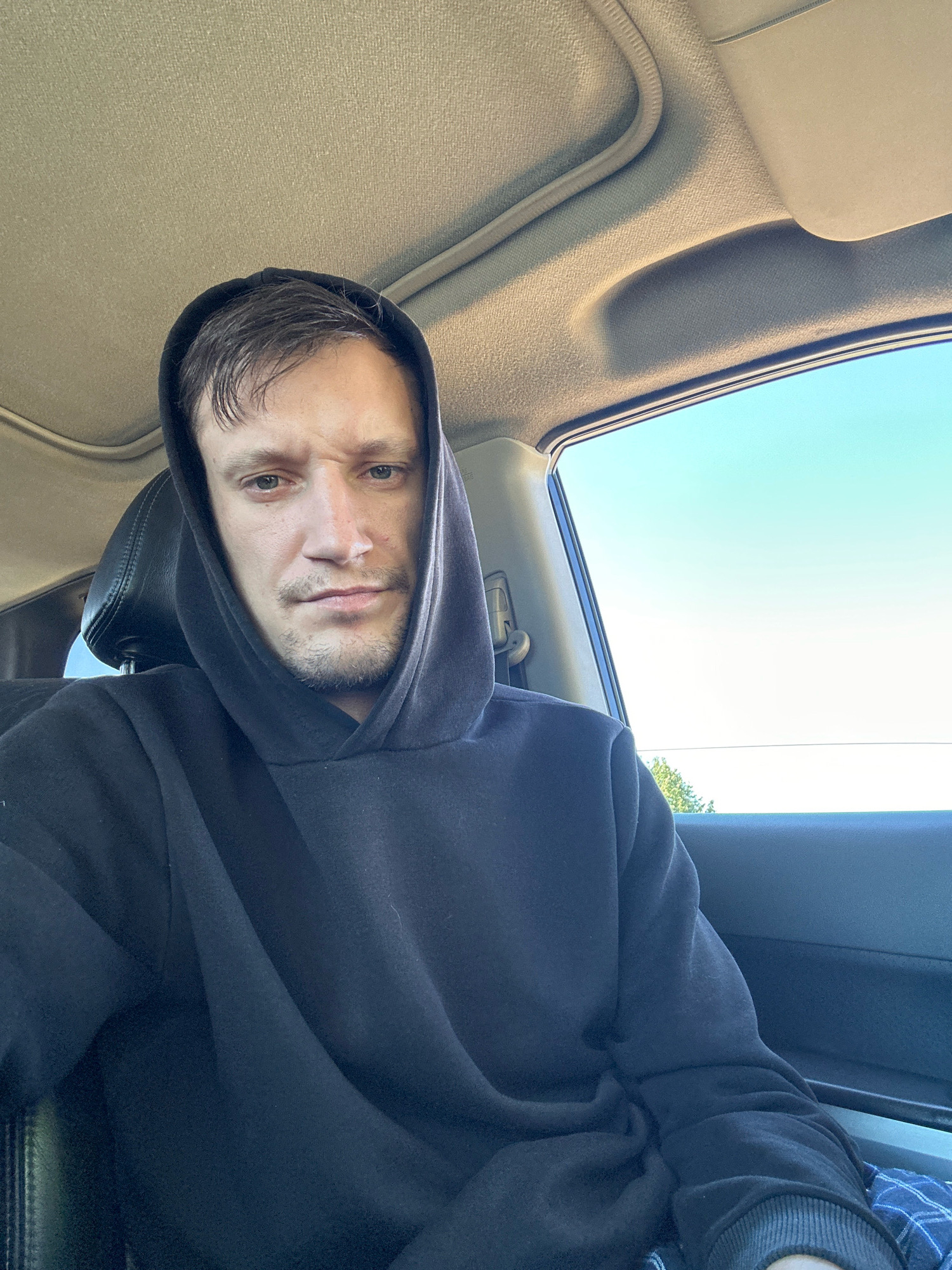 A man wearing a black hooded sweatshirt is sitting in the driver's seat of a car. He is looking directly at the camera with a neutral expression. The car's interior is visible, including the headliner and a portion of the passenger seat. Outside the car, the sky is clear and blue. The image appears to be a selfie taken during the daytime. The location in the city of Makiv, Ukraine, is not determinable from the provided image.