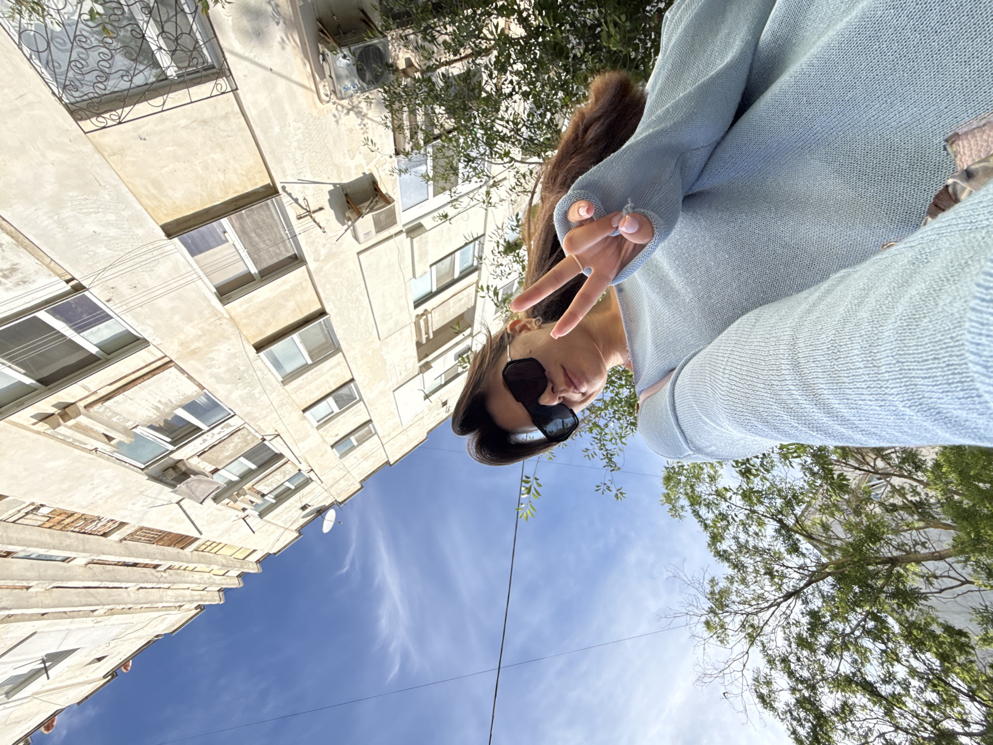 An eye-level, medium outdoor shot shows a young woman in a light blue sweater and black sunglasses taking a selfie. She is holding up two fingers in a peace sign. In the background, a tall, light-colored apartment building with many windows looms against a bright blue sky with wispy white clouds. Green trees are visible in the foreground. The camera is angled upwards.