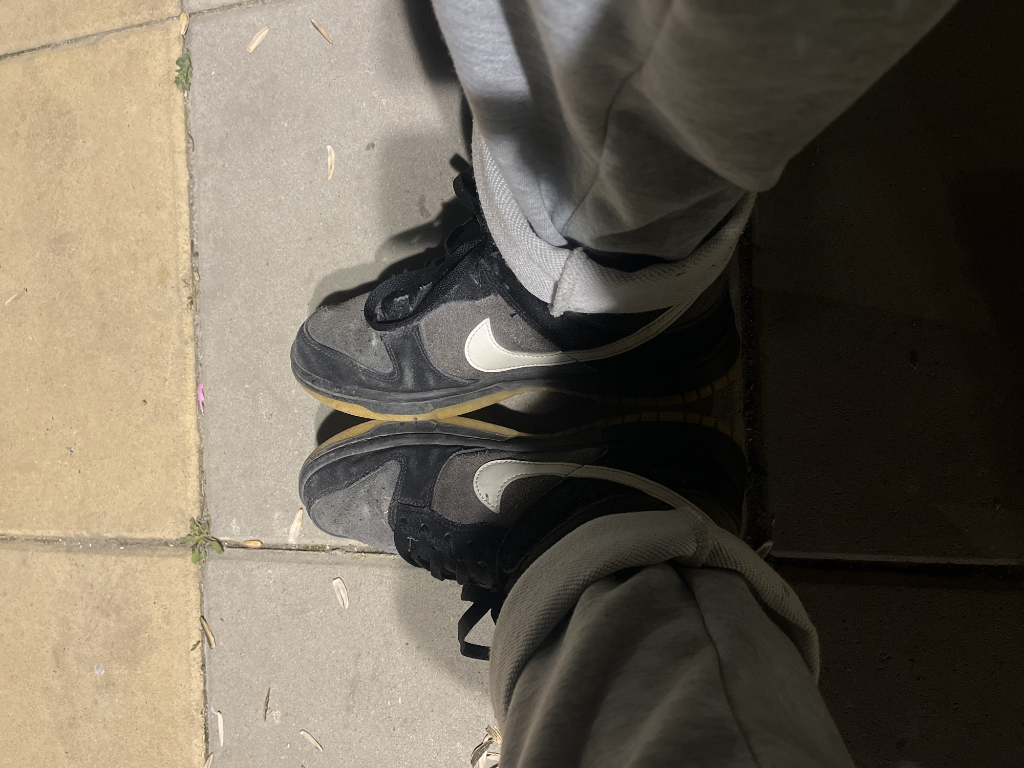 The image shows a person standing on a paved surface, likely a sidewalk. The surface is composed of square paving stones of two different colors - tan and gray. There is a small patch of greenery growing in the crack between the stones. The person is wearing gray sweatpants and black and gray Nike shoes with a white swoosh. The shoes are visibly worn. The location is unknown, so I cannot confirm if the location is Lozarevo, Bulgaria.