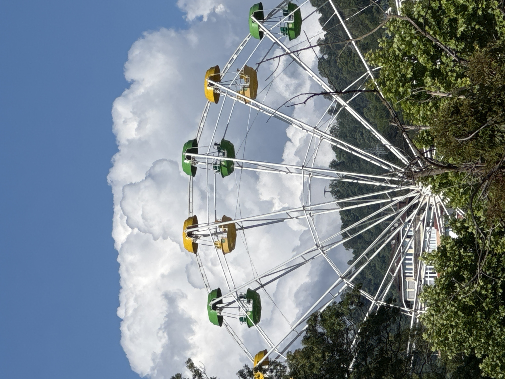 The image depicts a Ferris wheel against a backdrop of a blue sky with large, white cumulus clouds and a distant, tree-covered mountain range. The Ferris wheel has a white, spoke-like structure with numerous cabins attached. Some cabins are yellow-topped, and others are green-topped. The Ferris wheel is positioned in front of a building with white-framed windows, which is nestled amongst lush green trees and foliage. The trees in the foreground are dense and leafy, obscuring parts of the Ferris wheel's base. The lighting suggests it is daytime, with the sun casting shadows. The scene appears to be an outdoor amusement park or recreational area. This Ferris wheel is located in the Riviera Park in Sochi, Russia.