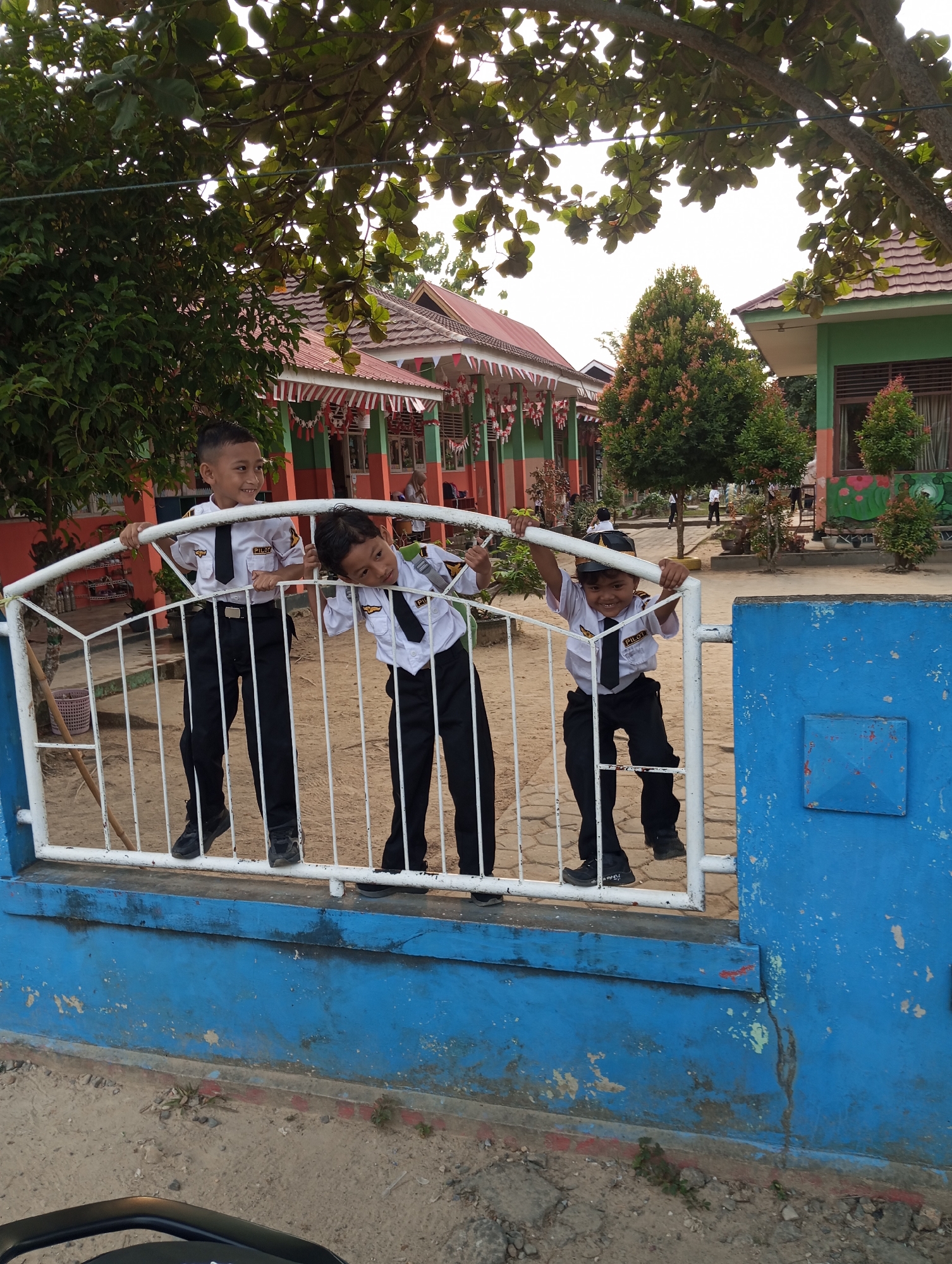 Three young boys in pilot uniforms, including white shirts with badges, black ties, and black pants, are leaning on a white metal gate in front of a school building. The boy on the left is standing straight and smiling, while the other two are leaning on the gate with playful expressions. Behind them, the school building has a red-tiled roof and green and orange walls, adorned with festive red, white, and blue banners. Lush green trees and bushes surround the school. The ground is a mix of dirt and pavement. This scene appears to be captured in front of a school in Pematang Reba, Indonesia.