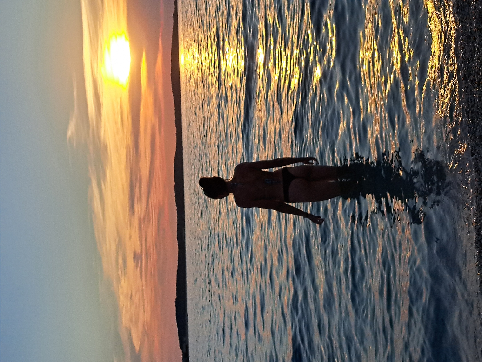 A person in a bikini stands in the water, facing the sunset. The sun is low on the horizon, casting a warm glow on the clouds and the water. The sky is a mix of orange, pink, and blue. The water is calm, with gentle waves reflecting the colors of the sunset. The person's silhouette is visible against the bright sky. The scene evokes a sense of peace and tranquility. There is no visible text in the image.
