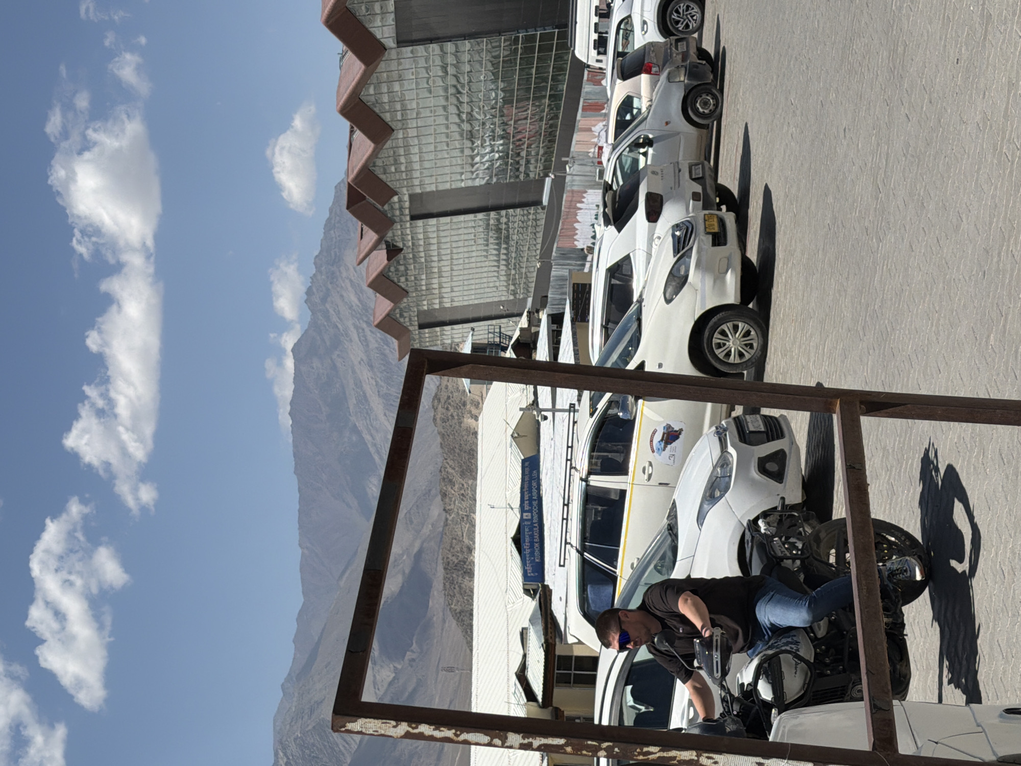 In Leh, India, a man is seated on a white and black motorcycle in the foreground, facing left, his right arm resting on the handlebars. He is wearing a dark t-shirt, blue jeans, sunglasses, and sneakers. The motorcycle is parked in front of a row of white cars. Behind the cars, a modern building with a glass facade and a distinctive zig-zag roofline is visible. Further in the background, a mountain range under a blue sky with scattered white clouds forms the backdrop. A sign is visible on the building that reads "KUSHOK BAKULA RINPOCHE AIRPORT, LEH". The ground is paved with grey interlocking bricks. A rusty metal frame is in the immediate foreground, partially obscuring the view of the man and motorcycle.