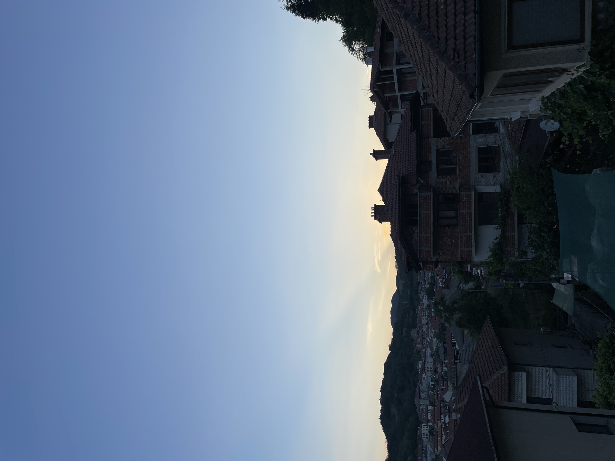 The image is a vertical, high-angle shot of a residential area in Zlatograd, Bulgaria, during twilight. The sky is a gradient of light blue, transitioning to a pale yellow near the horizon, with subtle sun rays visible. Below the sky is a panorama of houses with red-tiled roofs, nestled against a backdrop of green, tree-covered hills. The houses are a mix of completed structures and those under construction, characterized by brickwork and visible framing. Some of the houses have multiple stories and chimneys. Trees and vegetation are interspersed throughout the scene, with denser foliage on the hills. The foreground features the tops of several houses, with one on the far right displaying a light-colored facade and a window. Another house on the left has a dark roof and a section of white brickwork. The overall impression is of a quiet, developing town at dusk.
