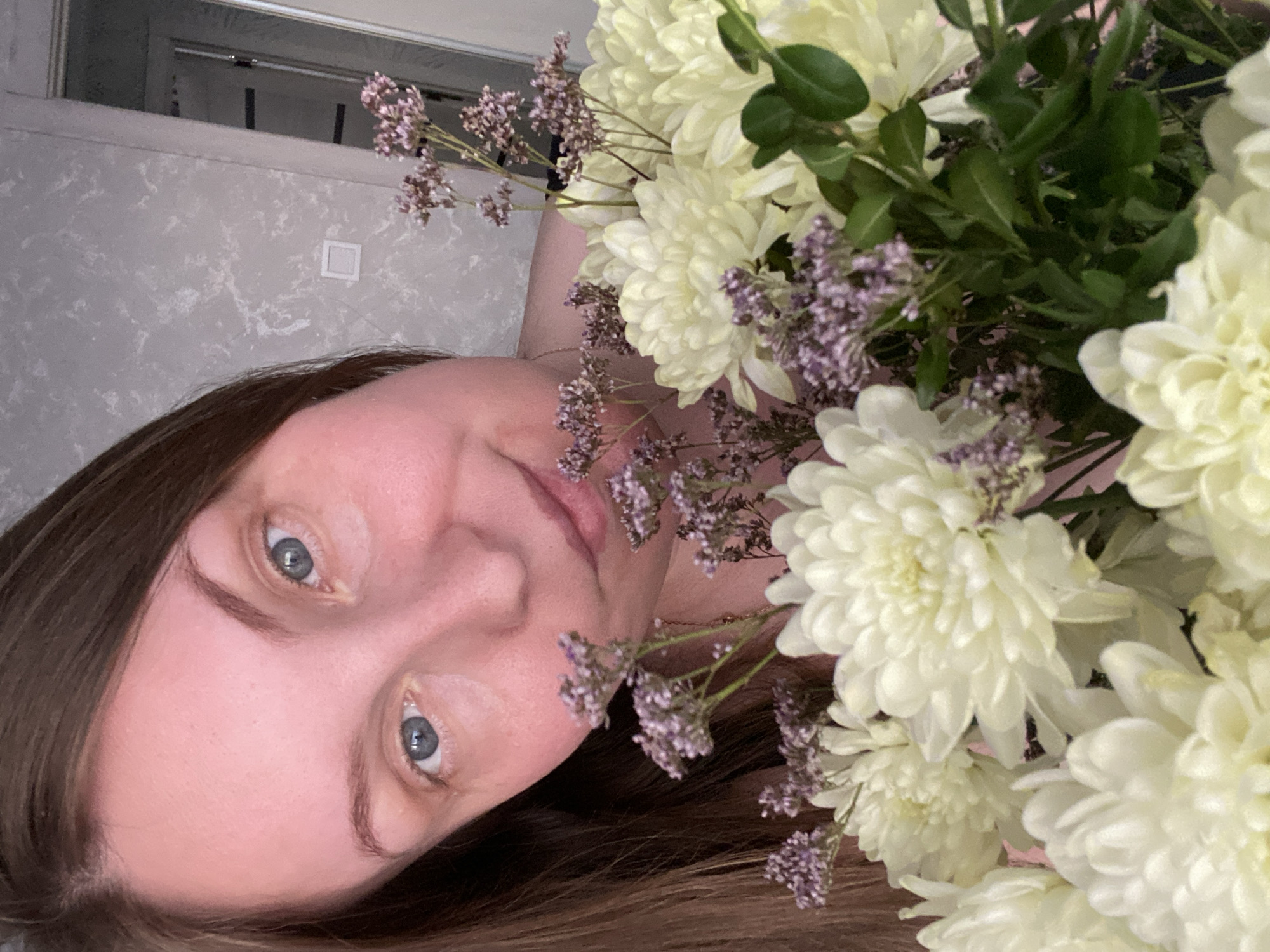 This image presents a close-up, tilted portrait of a woman's face alongside a vibrant bouquet of flowers, captured indoors in Noviy, Russia. The woman, positioned on the left side of the frame, has fair skin, long brown hair, and light blue or grey eyes, looking directly at the camera with a calm, neutral expression. Her face shows minimal or no visible makeup.
To her right, a large bouquet occupies the majority of the frame. It predominantly features white chrysanthemums, characterized by their full, fluffy petals. Interspersed among the white flowers are clusters of smaller, delicate light purple flowers, resembling statice or similar dried floral elements, and some dark green foliage.
The background behind the woman reveals a light-toned wall with a subtle, textured pattern that appears mottled or marbled. In the upper part of the background, a strip of a white door frame or window trim is visible, along with a small, white square object on the wall, likely a light switch or outlet, though no text is discernible. The lighting appears soft and even, suggesting an indoor setting that is well-lit, possibly by natural daylight or diffused artificial light. No specific time of day or weather cues are evident. The overall mood of the image is serene and pleasant.