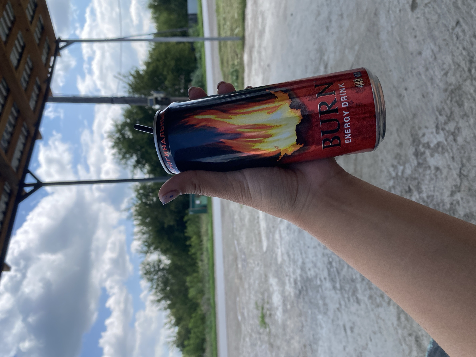 A person's hand holds a red can of "BURN ENERGY DRINK" with a flame design. The can has a black straw inserted. The background shows a cloudy blue sky, trees, and a road. The scene appears to be outdoors, possibly in a rural area or on the outskirts of a town like Антоньевка, Russia. The ground is paved with concrete or asphalt.