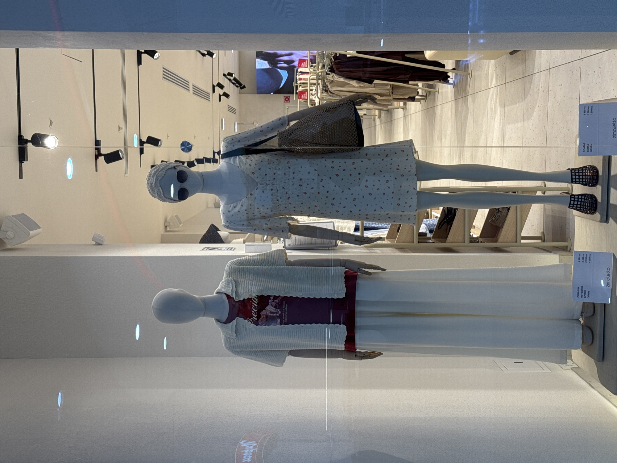 Here is a description of the image.
The image shows a store window in what appears to be a clothing store. There are two mannequins on display, one above the other. The top mannequin wears a short, light-colored dress with small floral patterns, a dark handbag over its shoulder, sunglasses, and a knitted hat. It also wears what looks like black strappy shoes.
The bottom mannequin is dressed in a long, white skirt and a white cardigan over a dark top with a design.
Reflections are visible on the glass window, reflecting the store's interior, including clothing racks and other merchandise. The overhead lighting is bright, with multiple spotlights.
The tags of the clothing are also visible.
The location is Unknown, Unknown.