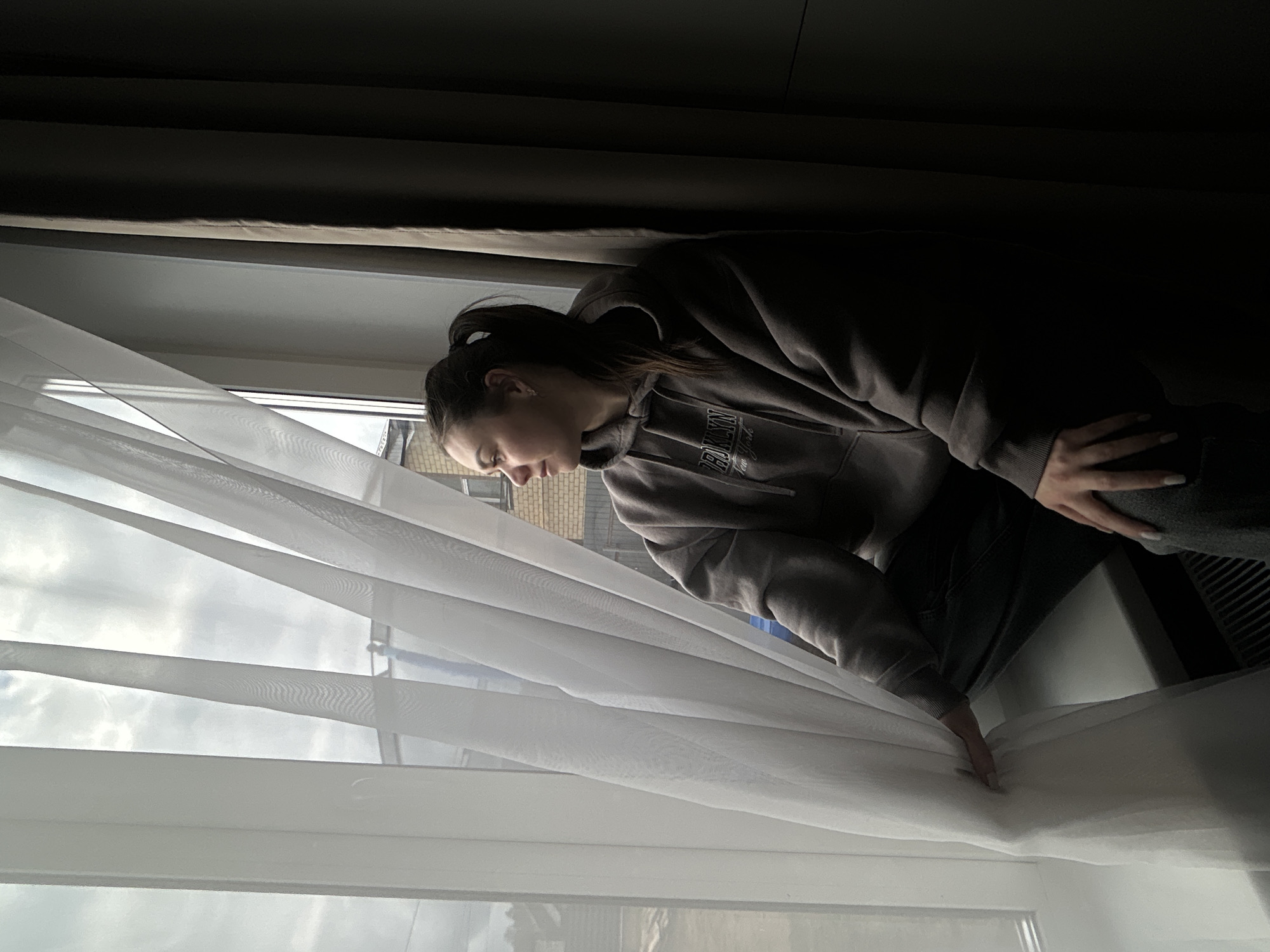The image, initially rotated, depicts a young woman looking out a window from a dimly lit indoor setting. She appears to be in her late teens or early twenties, seen in profile with dark hair tied back in a ponytail. She wears a dark brown or grey hooded sweatshirt with the text "BROOKLYN New York" partially visible across the front. Her hand, featuring long, light-colored nails, holds back a sheer white curtain to peer outside.
Beyond the window, a cloudy daytime sky is visible, along with parts of brick buildings, suggesting an urban or suburban environment. The light outside is bright but diffused, indicating an overcast day. Above the sheer white curtains, darker, heavier curtains or blinds are drawn back. Below the window, a dark-colored radiator or vent is partially visible. The woman's facial expression conveys a sense of contemplation or quiet curiosity as she gazes out, observing the scene in Chaikovsky, Russia. The stark contrast between the bright exterior and the dark interior of the room is a notable visual element.
