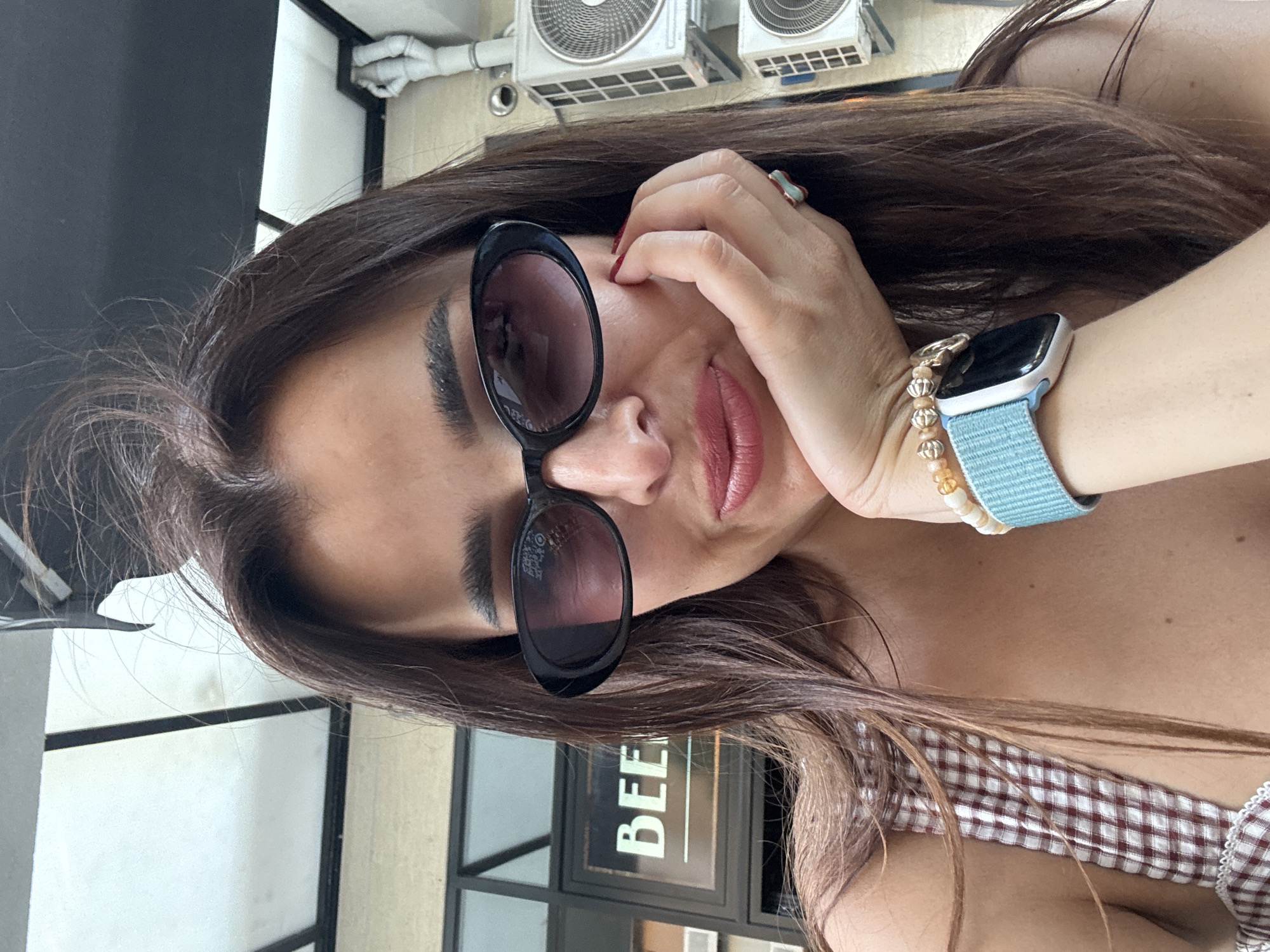 A woman is captured in a close-up, selfie-style photograph, resting her chin on her hand. She is wearing dark, oval-shaped sunglasses, and her lips are painted a muted pink. Her brown hair is long and slightly messy, framing her face. She has on a checked top and several bracelets on her wrist, including one with beads and another with an apple watch. In the background, a sign with the word "BEER" is visible through a window, suggesting an outdoor or semi-outdoor setting, possibly a restaurant or bar. Two air conditioning units are mounted on a wall to the right of the frame. The overall lighting suggests daytime.