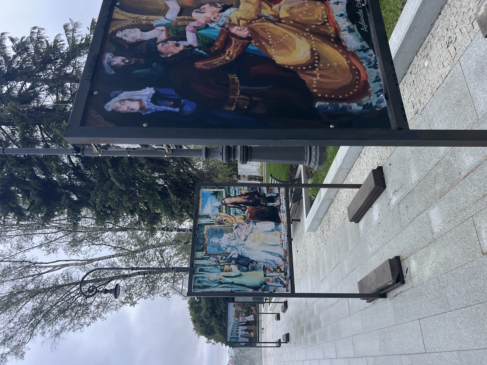 The image displays an outdoor exhibition of paintings mounted on metal stands along a paved walkway. The paintings appear to be figurative, with one prominently featuring a group of individuals in historical or theatrical attire. The closest painting is in the foreground, partially obscuring another painting to its left and another further back. The ground is paved with large grey granite tiles. In the background, trees and a lamppost are visible. The sky is overcast, suggesting a cloudy day. A signature is visible in the lower right corner of the closest painting.