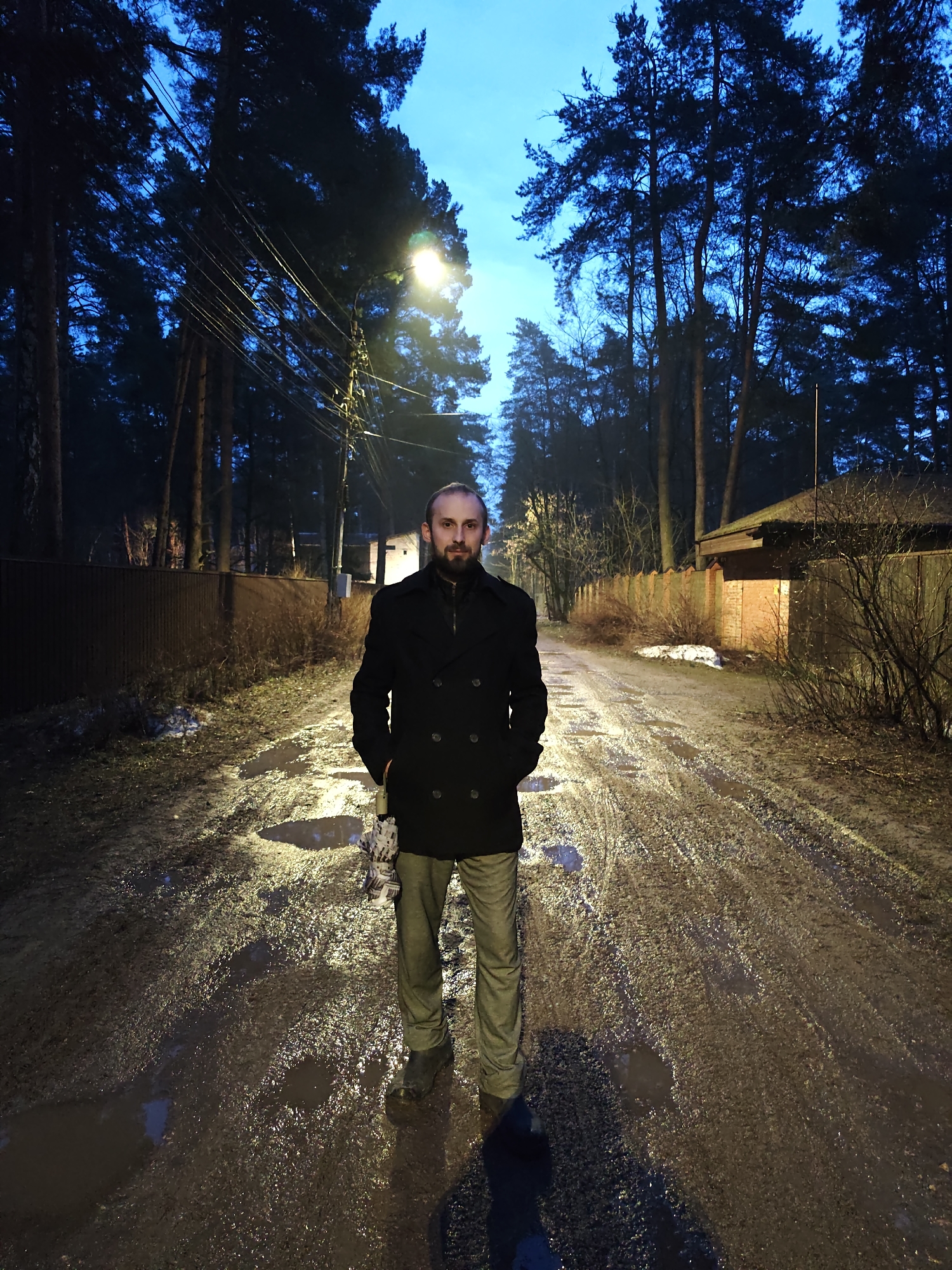 A full-length photograph depicts a bearded man standing in the center of a muddy, unpaved road lined with trees and fences. He is wearing a dark, double-breasted overcoat, light-colored pants, and dark shoes. His hands are in his pockets, and he is looking directly at the camera. Overhead street lighting illuminates the scene, creating a stark contrast between the light and shadows. Puddles of water reflect the light on the road's surface. The background includes trees, wooden fences, and a glimpse of a brick building, suggesting a residential area. This image could be from a location in Kratovo, Russia. The image portrays an individual standing amidst a somewhat rural or underdeveloped section of an urban area.