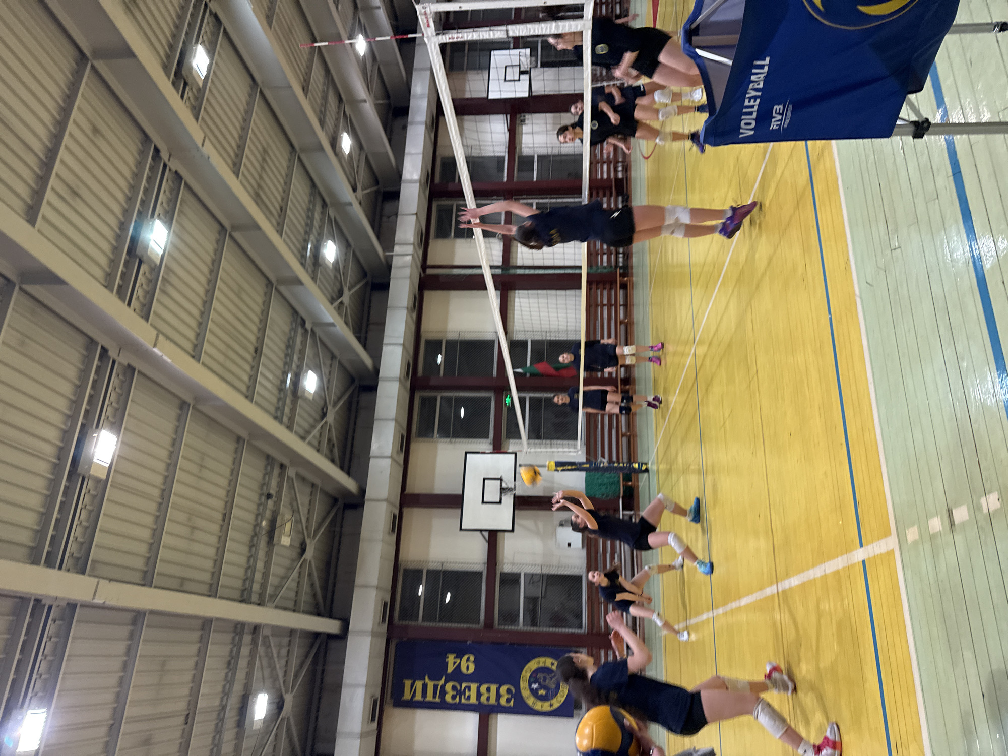 The image depicts a volleyball game in progress inside a gymnasium. Players, all female, are actively engaged in the game. The net is visible, with a volleyball in the air near the net. The background includes basketball hoops, a banner, windows, and the gymnasium's ceiling structure with lighting fixtures. A banner with "ЗВЕЗДИ 94" is seen in the background. The banner suggests that the location is likely in Sofia, Bulgaria, as "ЗВЕЗДИ 94" (Zvezdi 94) is a known Bulgarian volleyball club. There is a volleyball-branded object near the right side of the image. The volleyball court's colors are yellow and green.